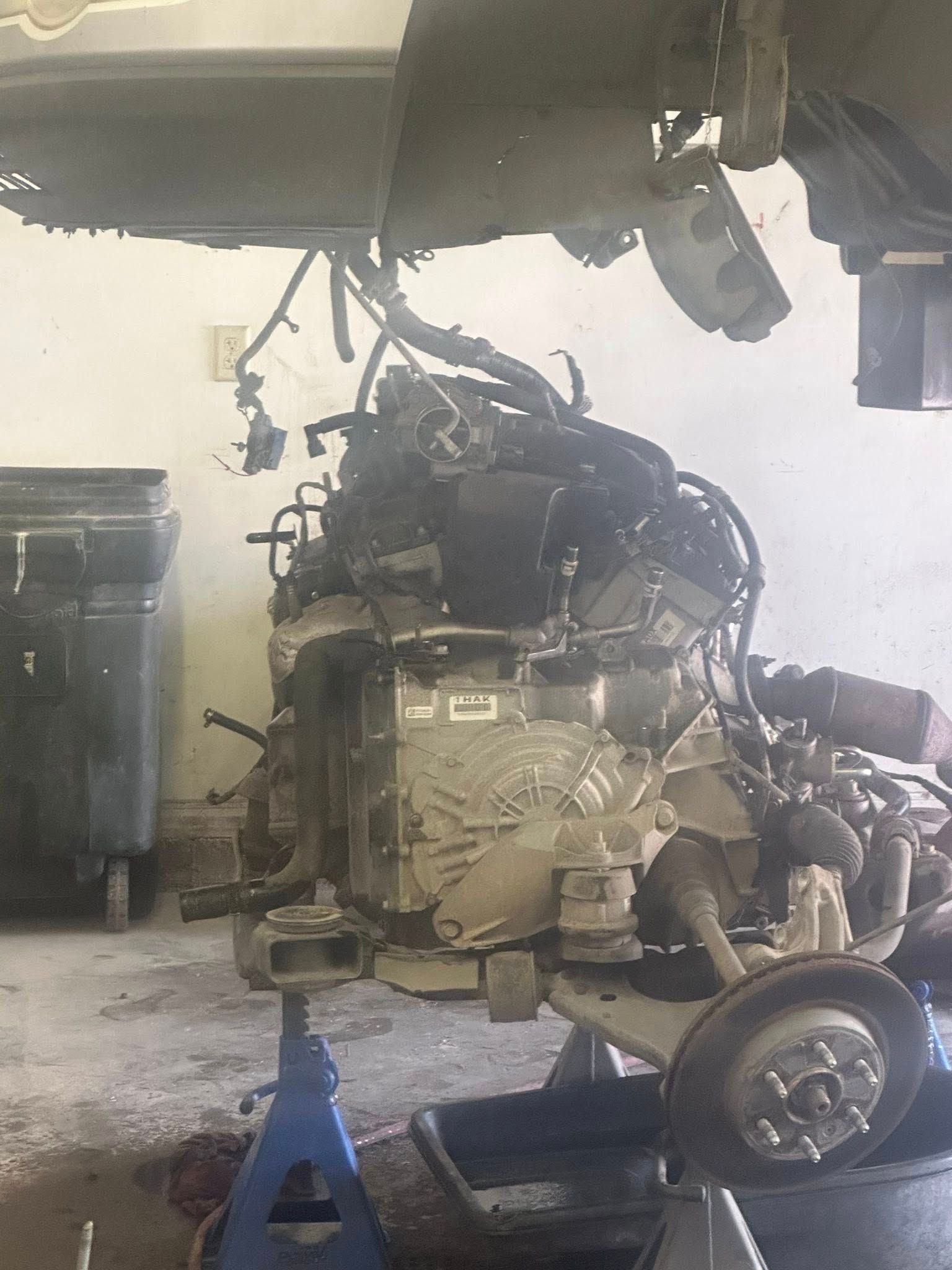 A car engine and transmission assembly suspended from an engine hoist inside a garage with a jack stand underneath.