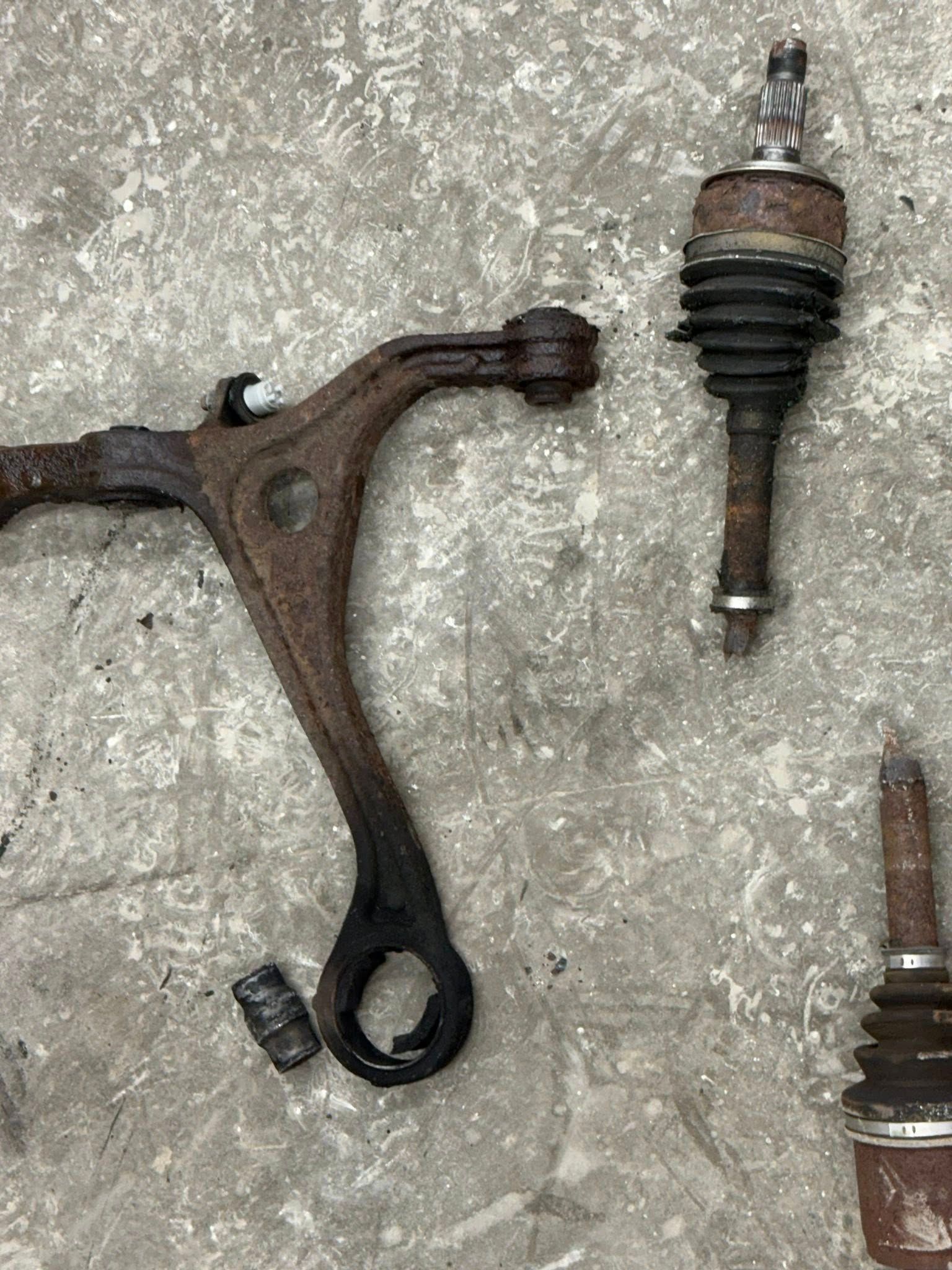 A rusted metal vehicle control arm and two suspension ball joints lying on a gray concrete surface.