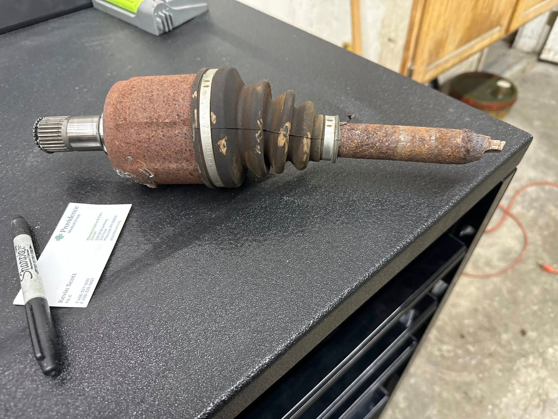 A heavily rusted automotive CV axle sits on a black workbench next to a marker and a business card.