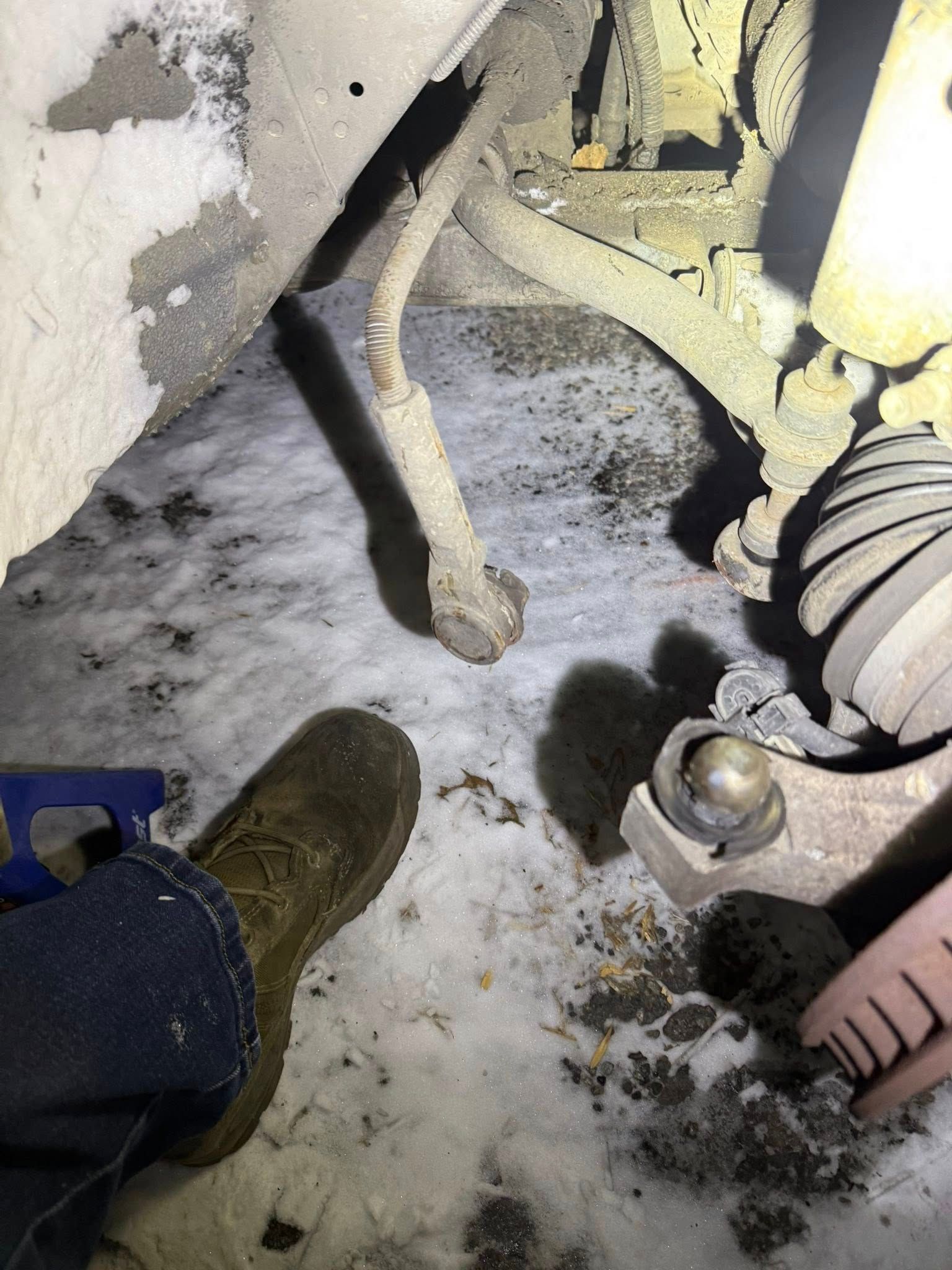 A person’s foot near a disconnected vehicle sway bar link and ball joint in a snowy area.