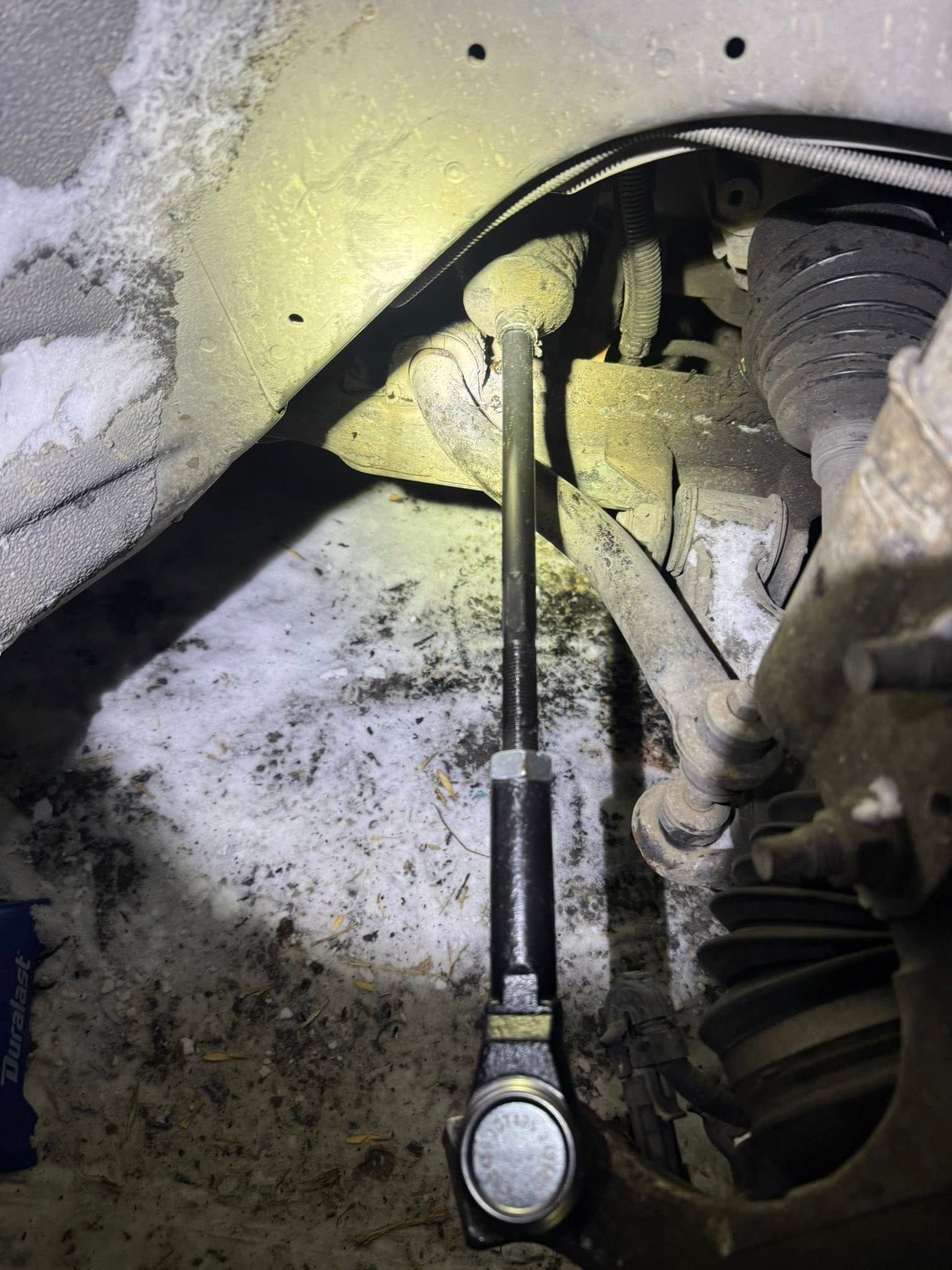 A newly installed, black sway bar link stands vertically in a dirty, snow-covered vehicle wheel well.