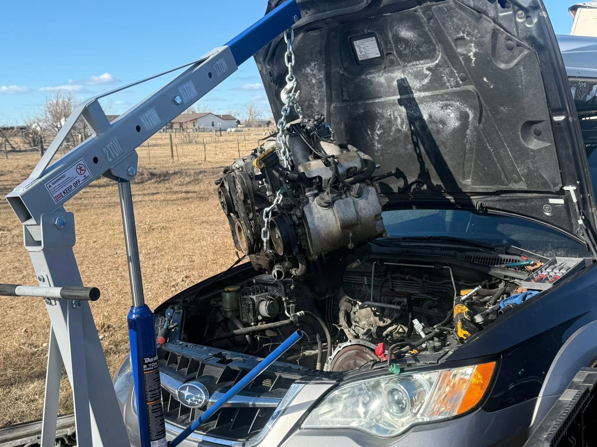 A gray engine hoist lifts a car engine out of the open hood of a dark sedan parked in a grassy, rural field.