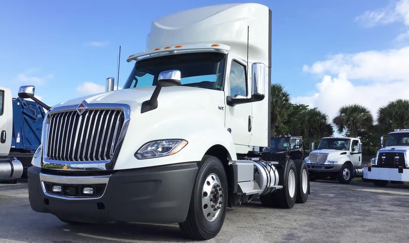 Blue Mack semi-truck driving on a road in a mountainous, green landscape under a cloudy sky.