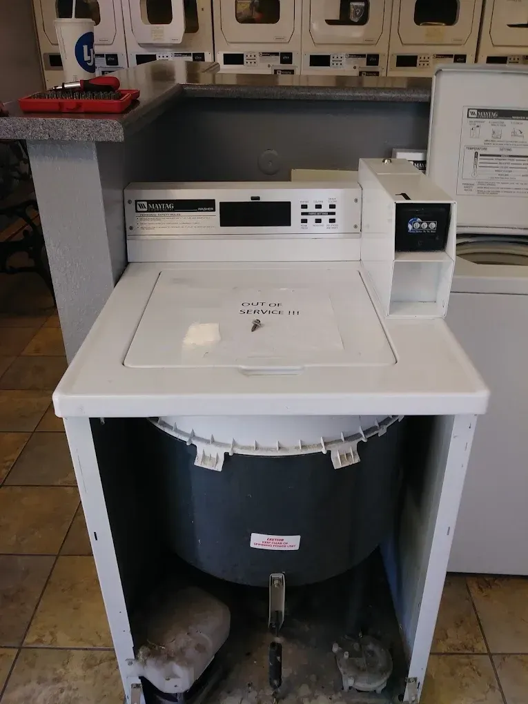 A top-loading washing machine in a laundromat, white and gray, coin-operated.