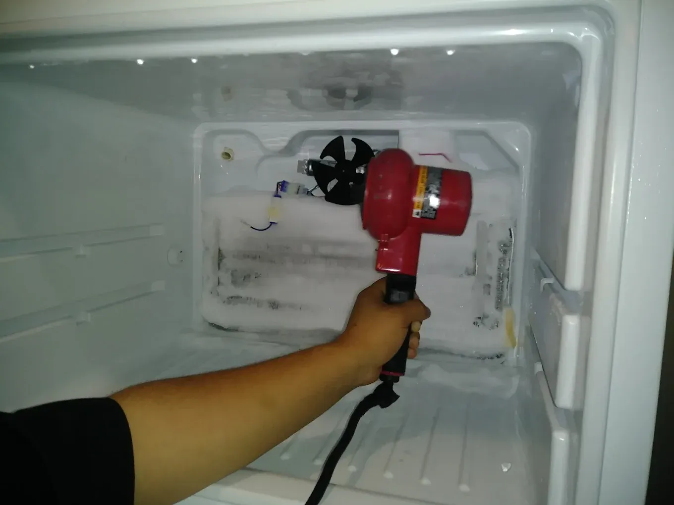 Person defrosting a freezer with a red heat gun. White interior, ice buildup, and a fan are visible.