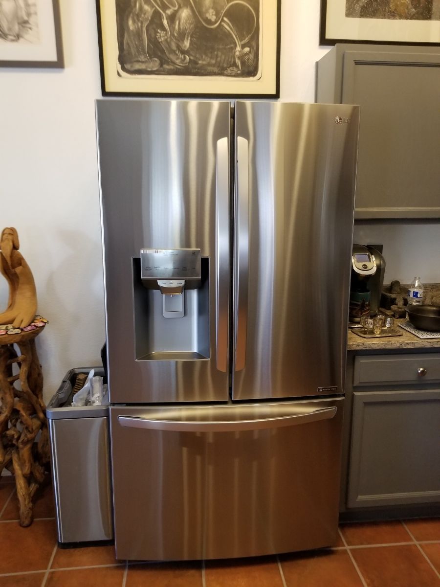 Stainless steel refrigerator with water dispenser, next to a trash can and cabinet.