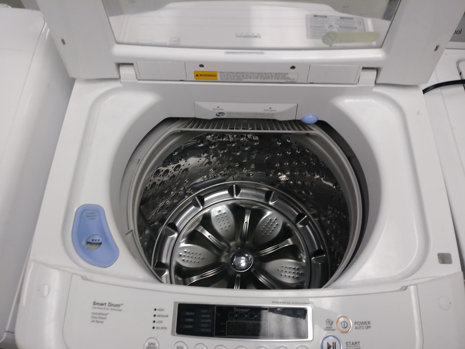 Top-load washing machine, open lid. Stainless steel drum with central impeller. White machine, blue button.