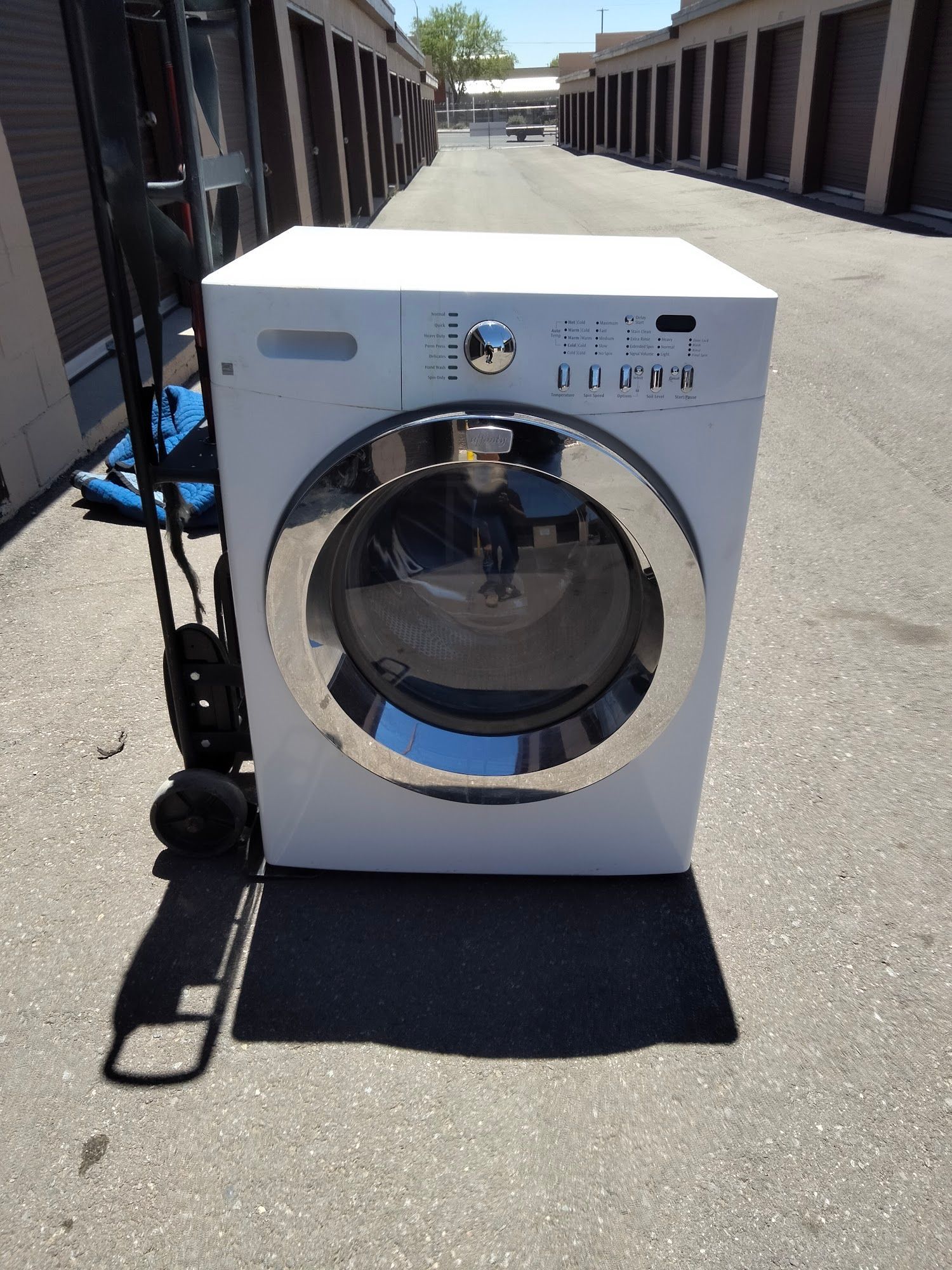 White front-load washing machine outside. Gray pavement with storage units in the background.