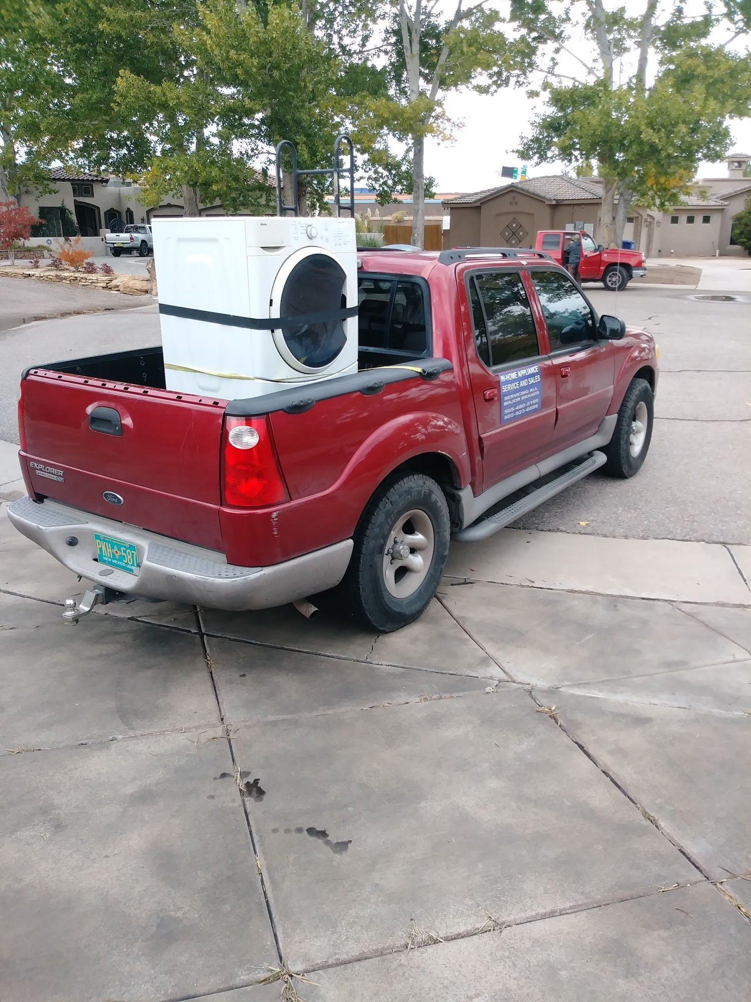 Red pickup truck carrying a white washing machine in its bed; parked on pavement.