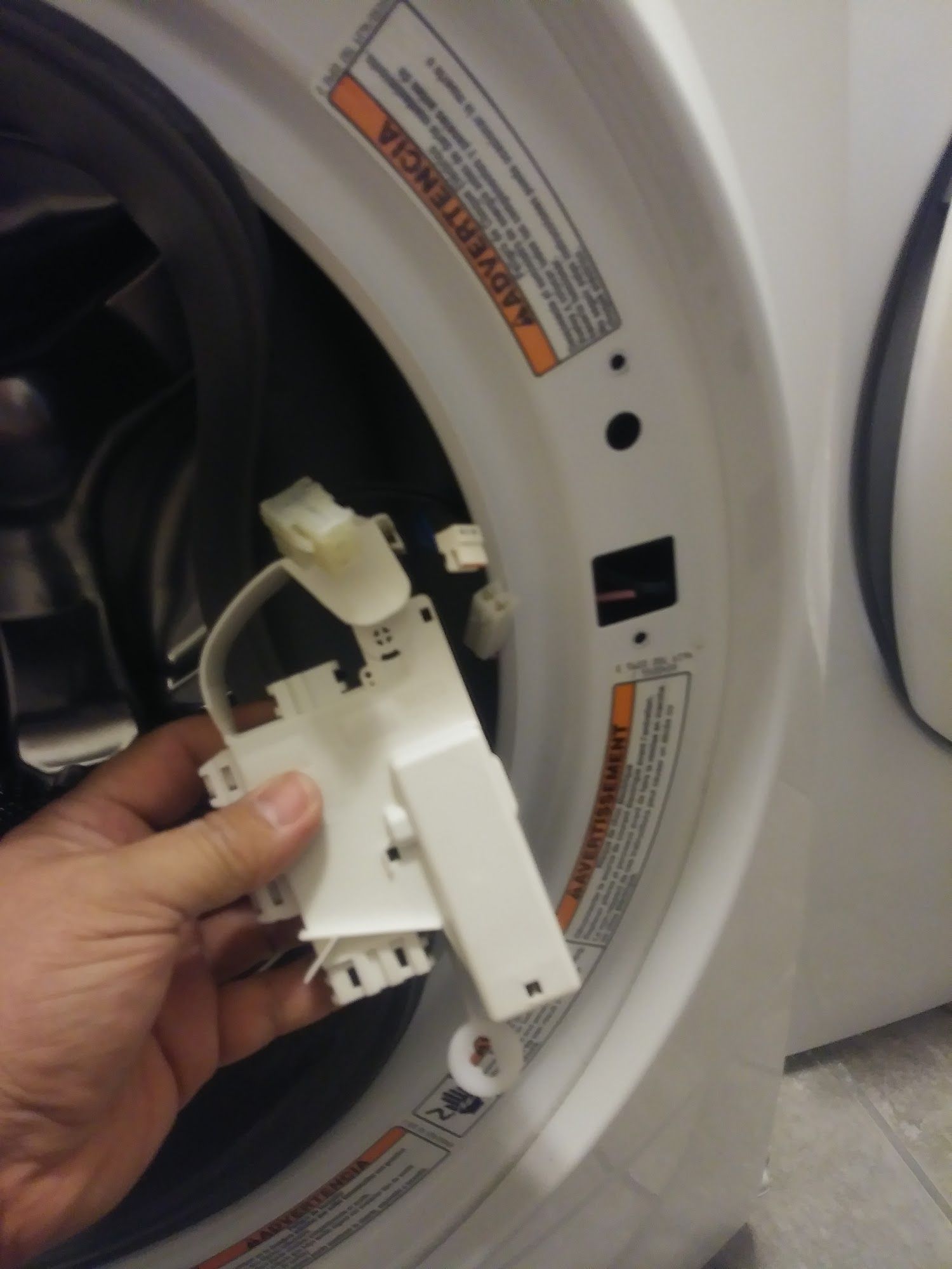 Hand holding a white washing machine door lock mechanism. The machine's door is open.
