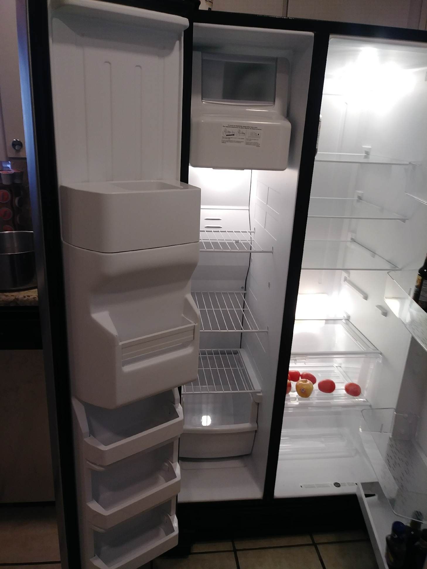 Open refrigerator, showing shelves, drawers, and ice maker. Tomatoes visible.