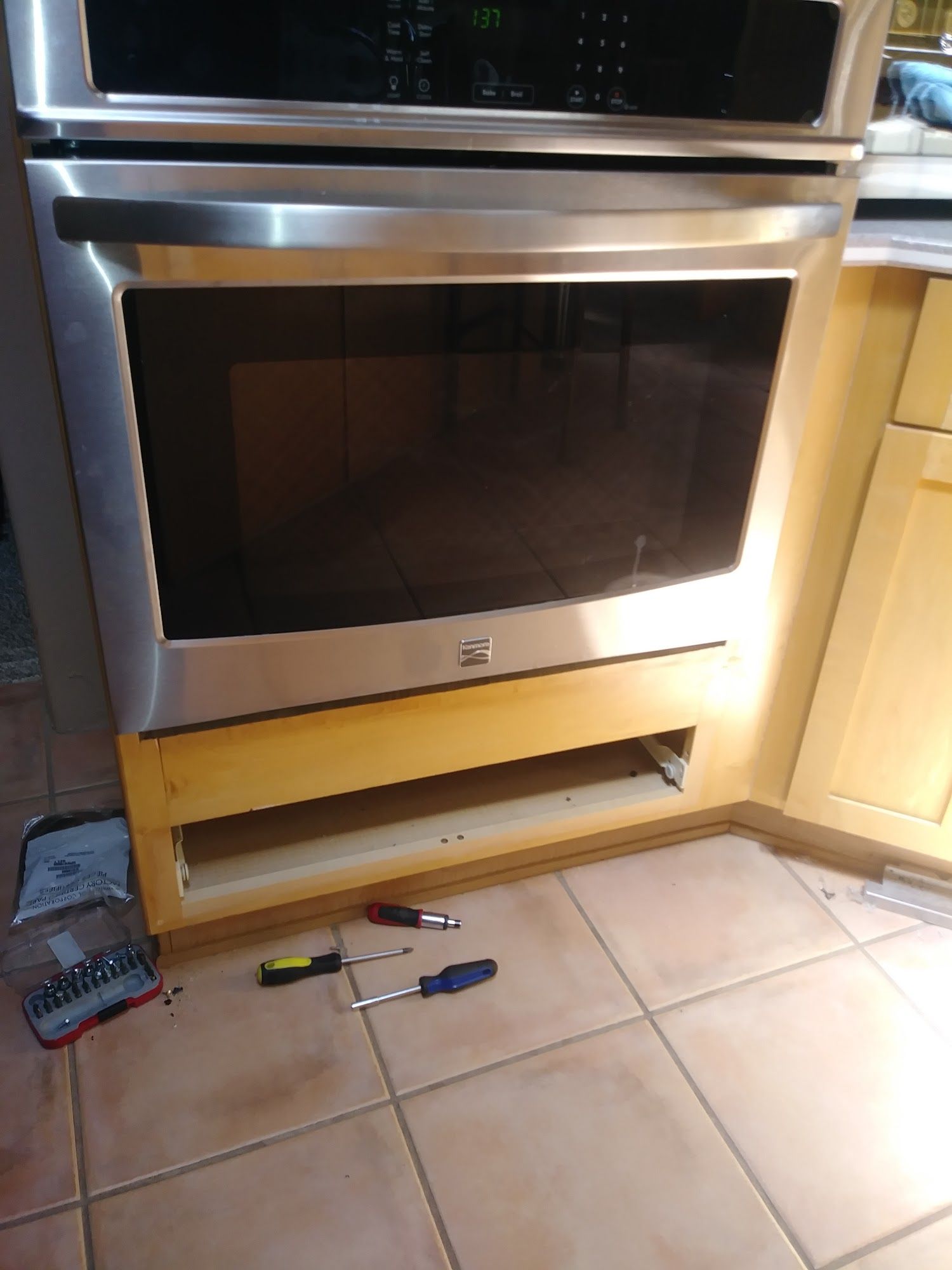 A stainless steel oven installed in a wooden cabinet. A lower drawer is removed, with tools on the floor.