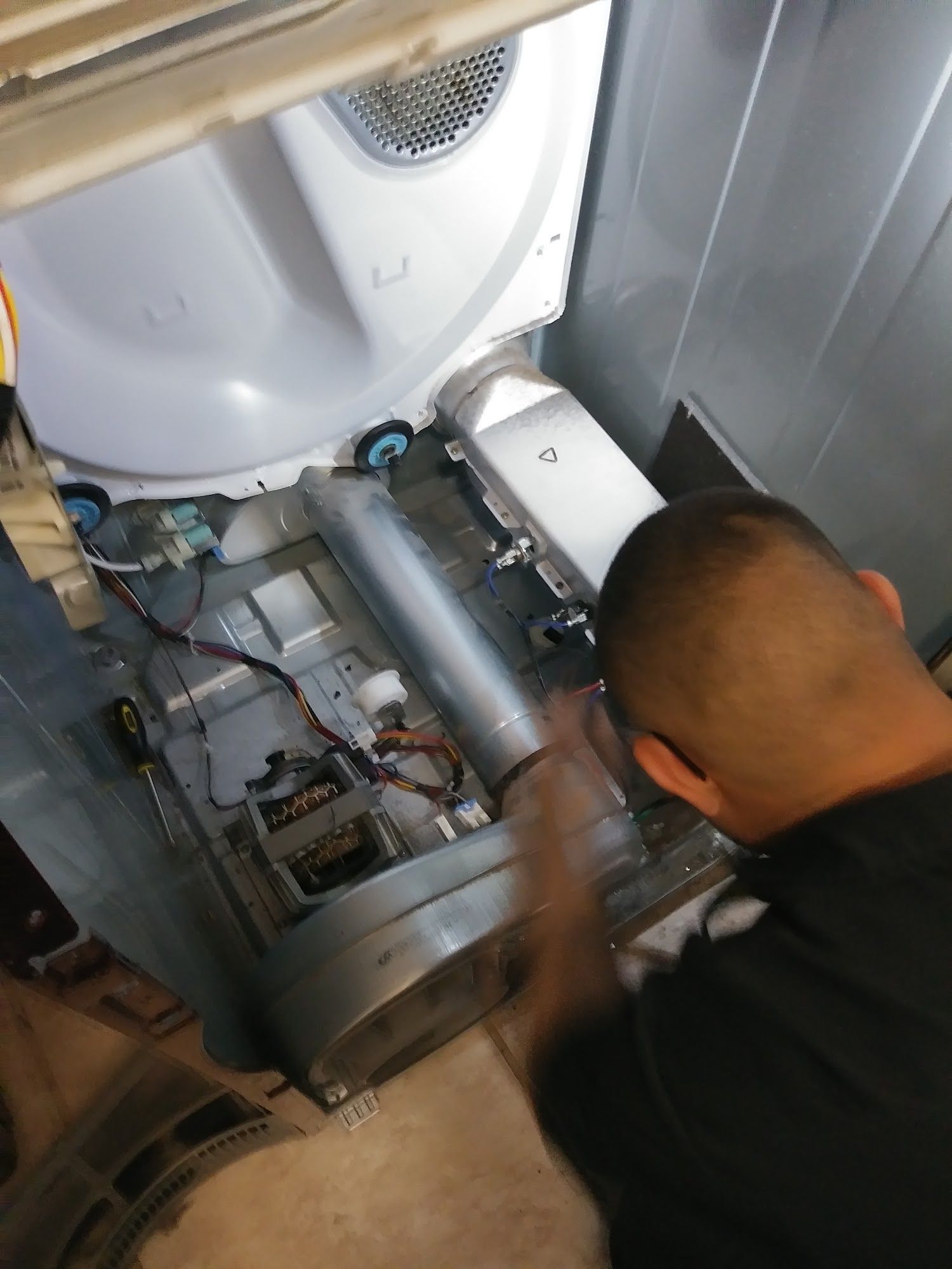 Man repairing the interior of a dryer, surrounded by parts. Gray and white appliance with visible components.