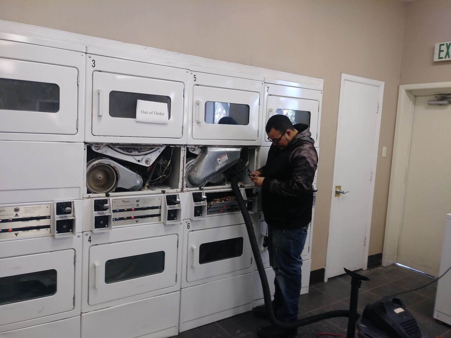 Man using a vacuum to clean a dryer in a laundromat. Dryers are white, the man is dark-haired, wearing jeans.