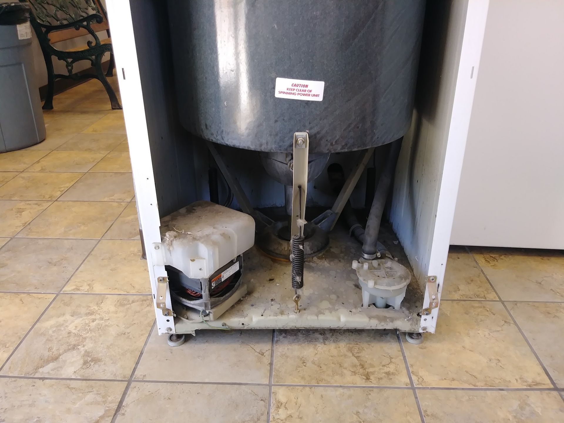 Backside of a white washing machine showing motor, tub, and connecting parts on a tiled floor.