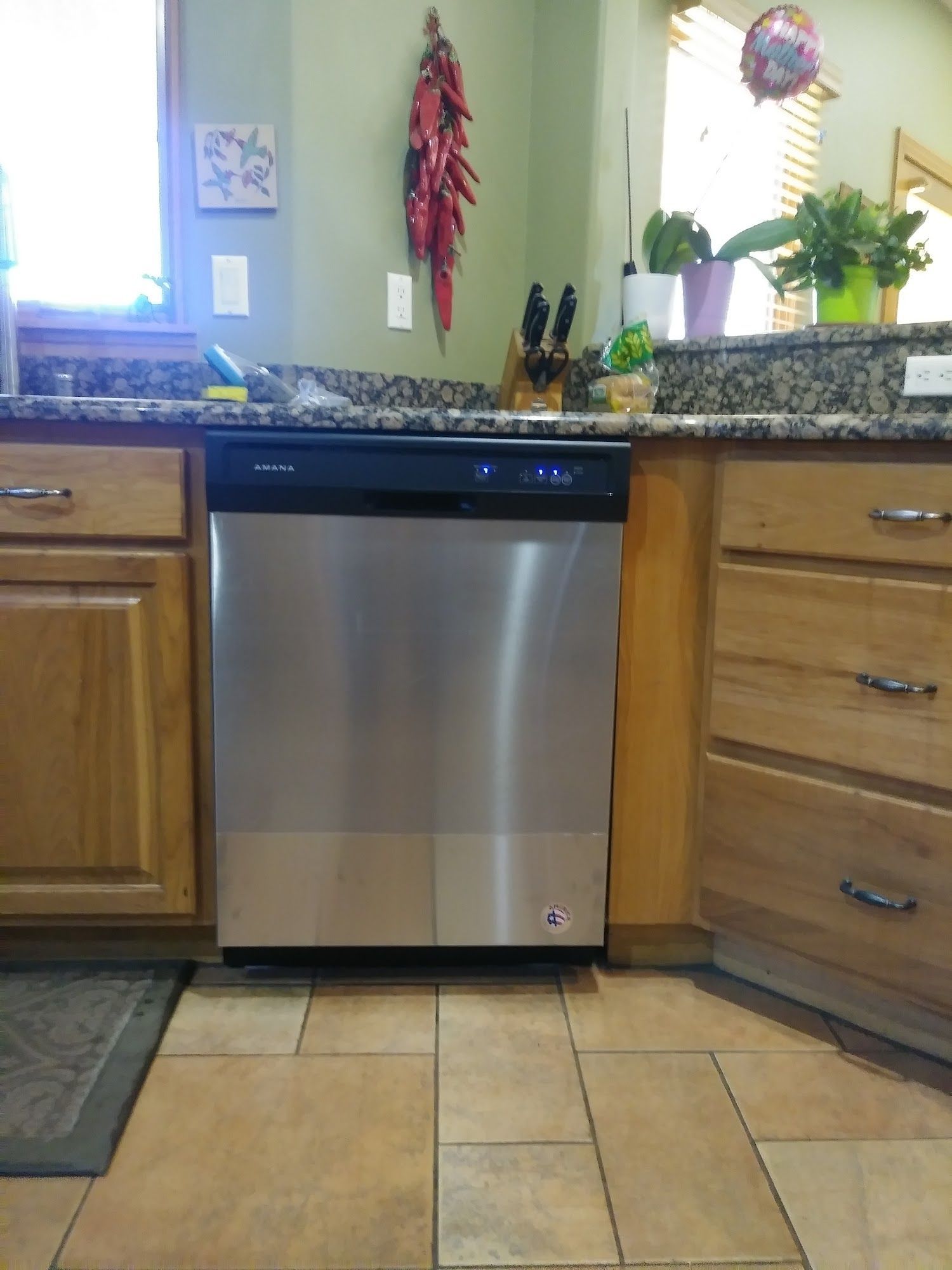 Stainless steel dishwasher in a kitchen with light wood cabinets, tile floor, and a hanging chili pepper decoration.