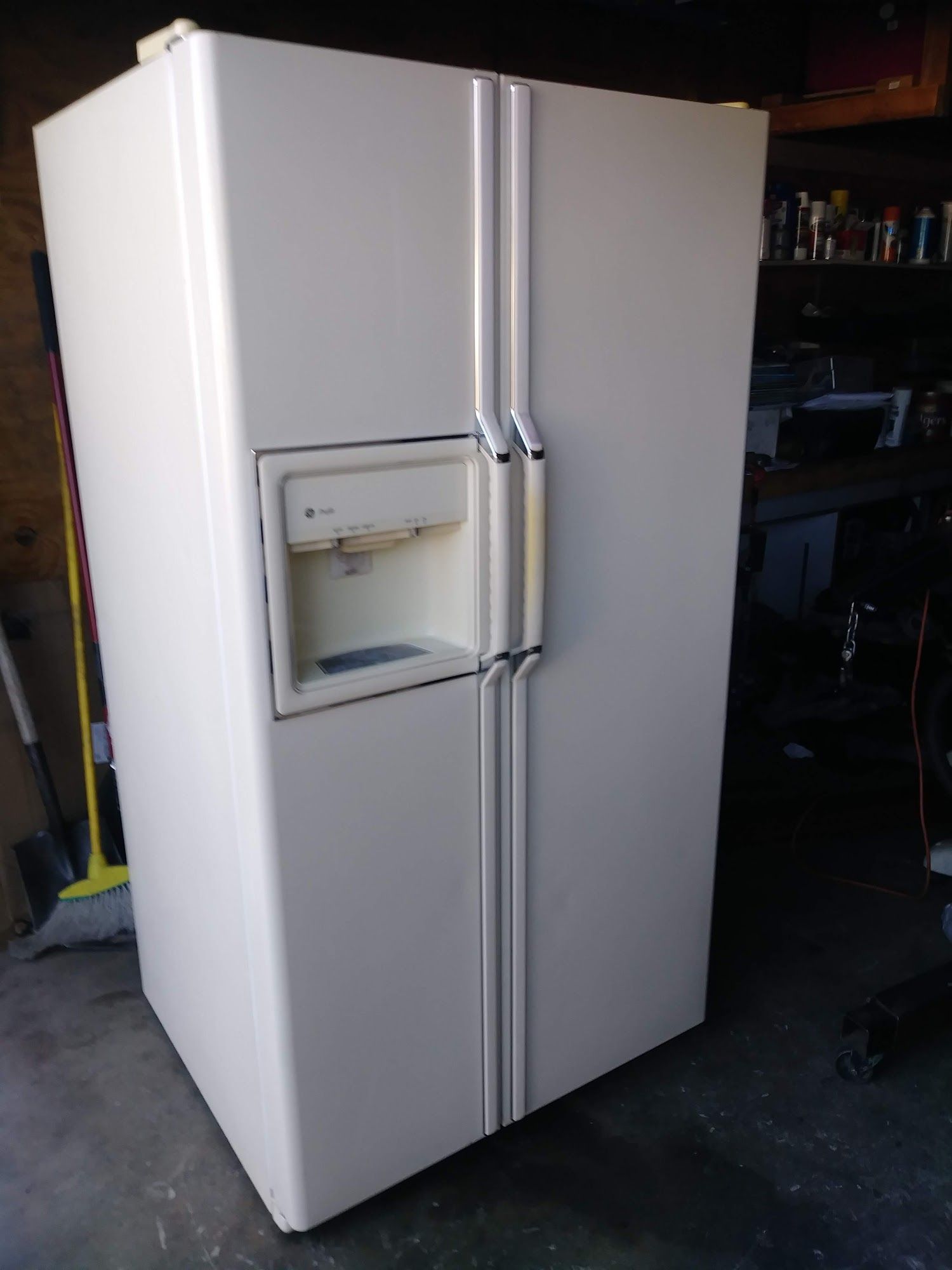Cream-colored side-by-side refrigerator with water dispenser and handles, set in a garage.