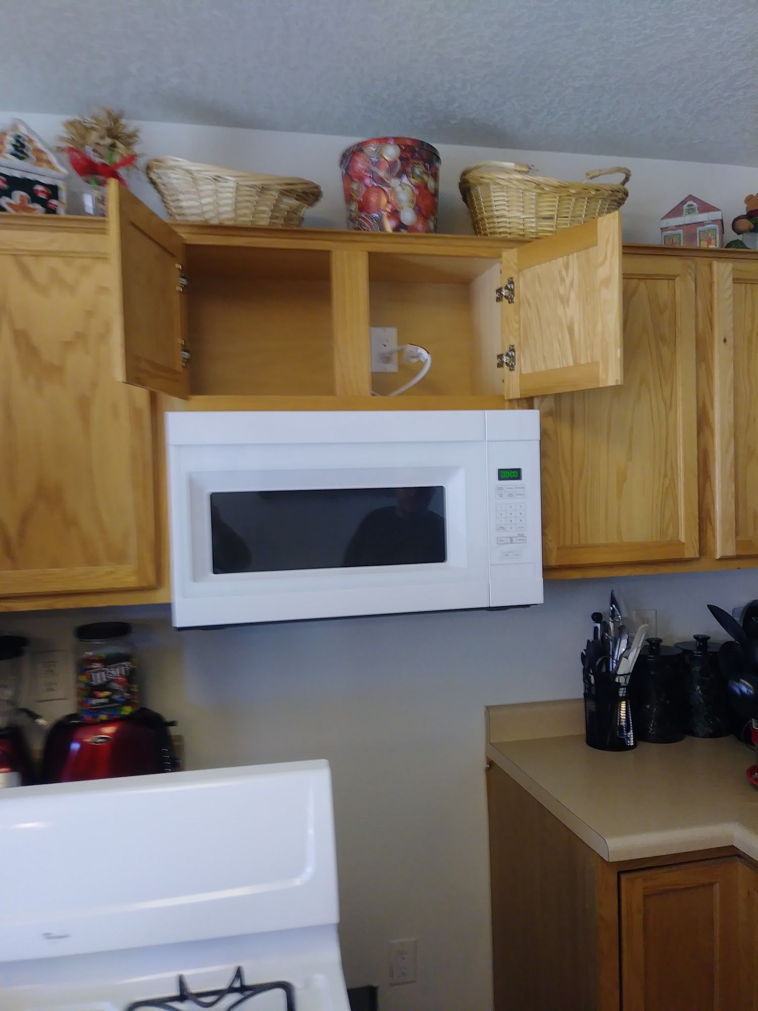 A microwave installed under a kitchen cabinet with open doors. Light wood cabinets, white microwave, baskets on top.