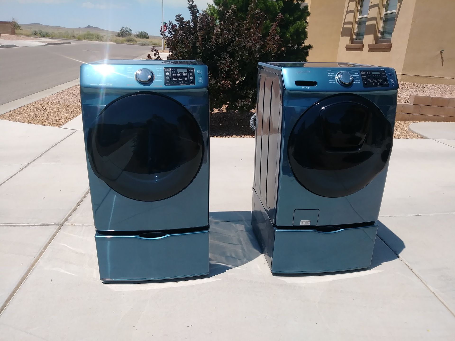 Two teal-colored Samsung washer and dryer units on pedestals, outdoors on a sunny day.