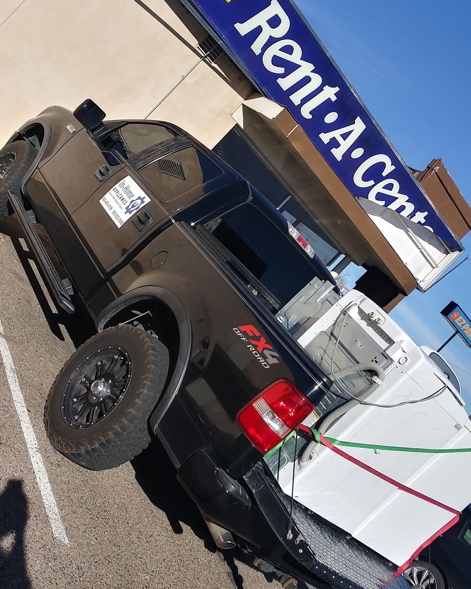 Black Ford pickup truck with washing machine in bed, parked outside a Rent-A-Center.