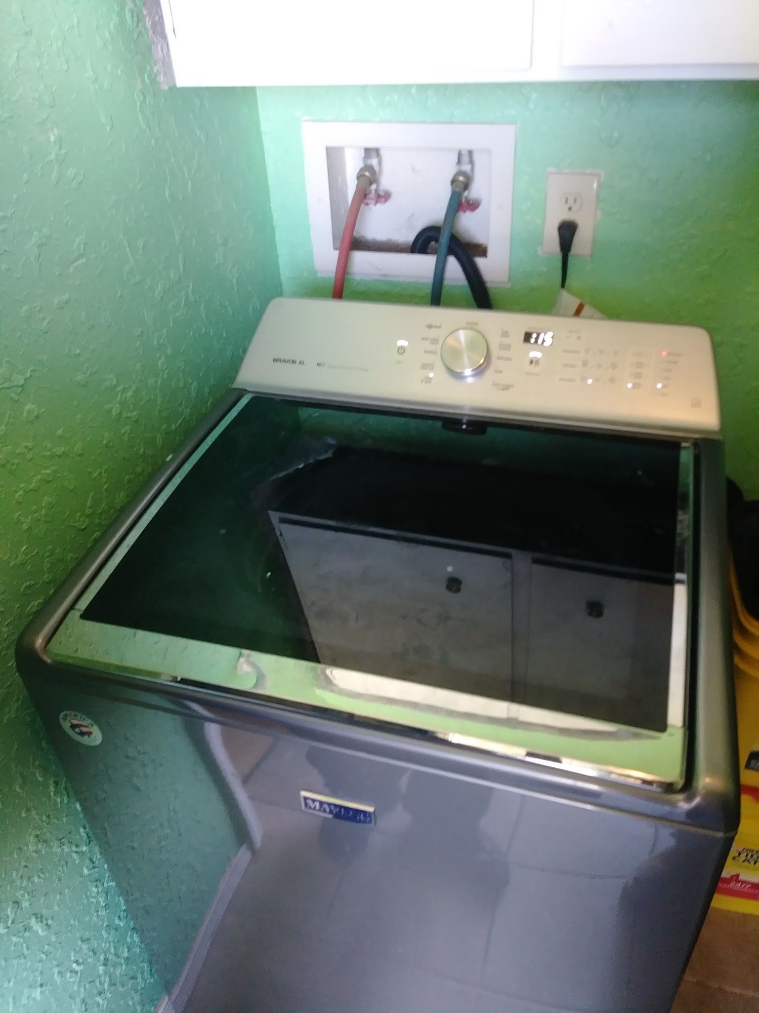 Maytag top-load washing machine against a green wall; water hookups and outlet visible.