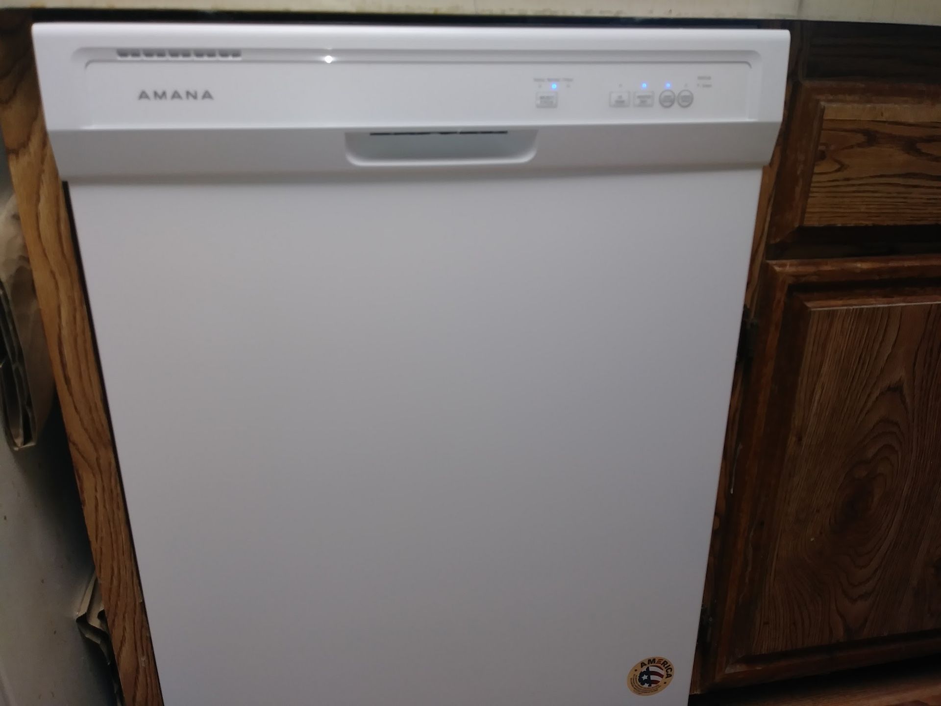 White Amana dishwasher in a kitchen, nestled between wooden cabinets.