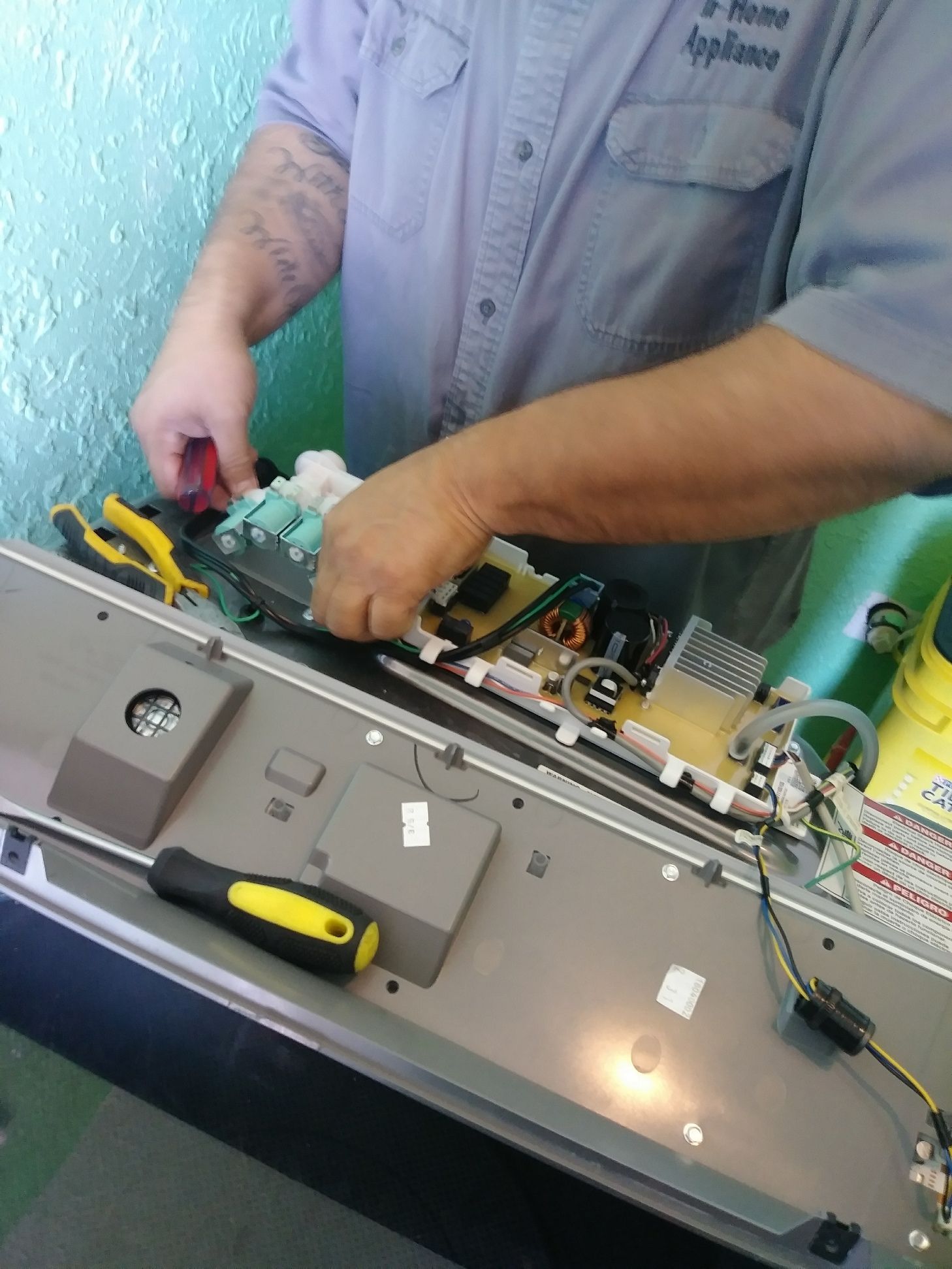 Man repairing appliance; hands focused on circuit board, using tools. Light blue shirt, in a brightly lit space.