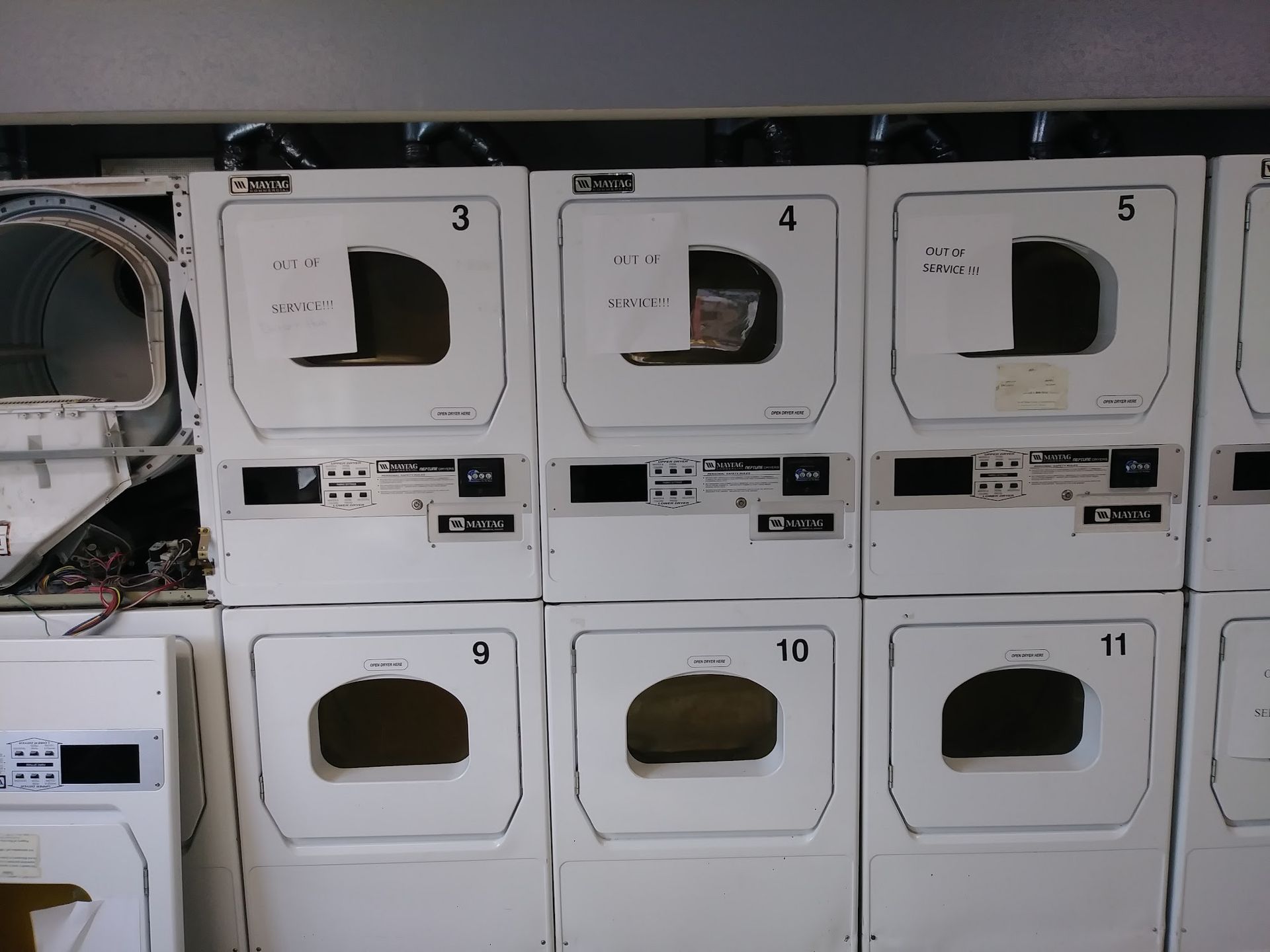 Row of white dryers in a laundromat; one is missing its door and interior components. Numbers 3-5, 9-11 visible.