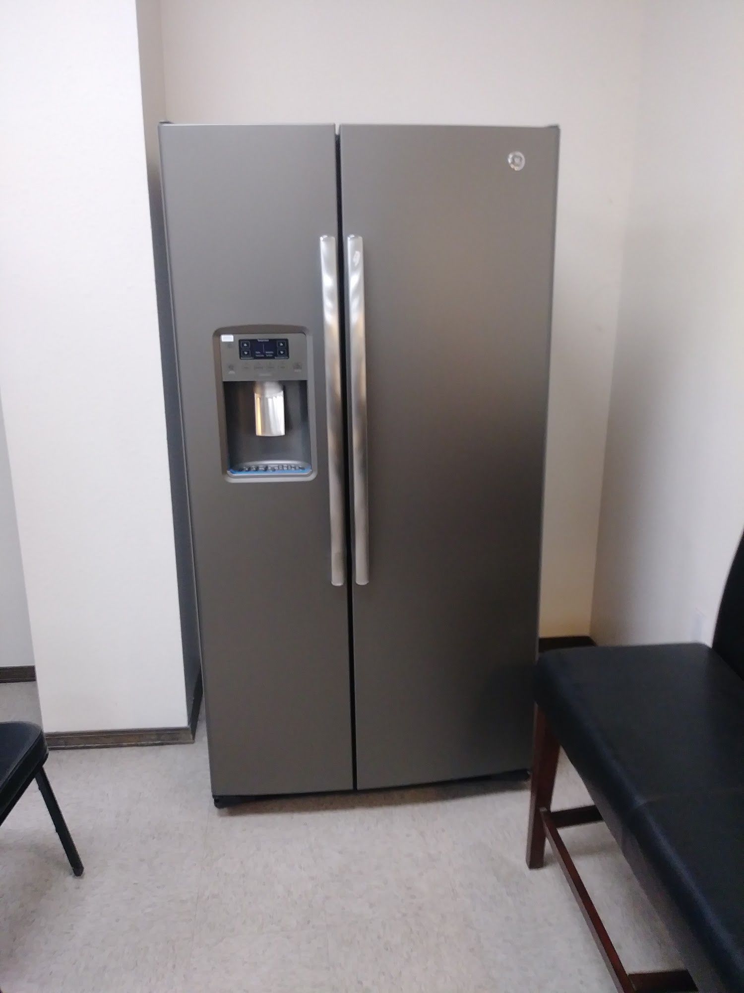 Side-by-side stainless steel refrigerator with water dispenser. Located in a room with chairs and light-colored flooring.