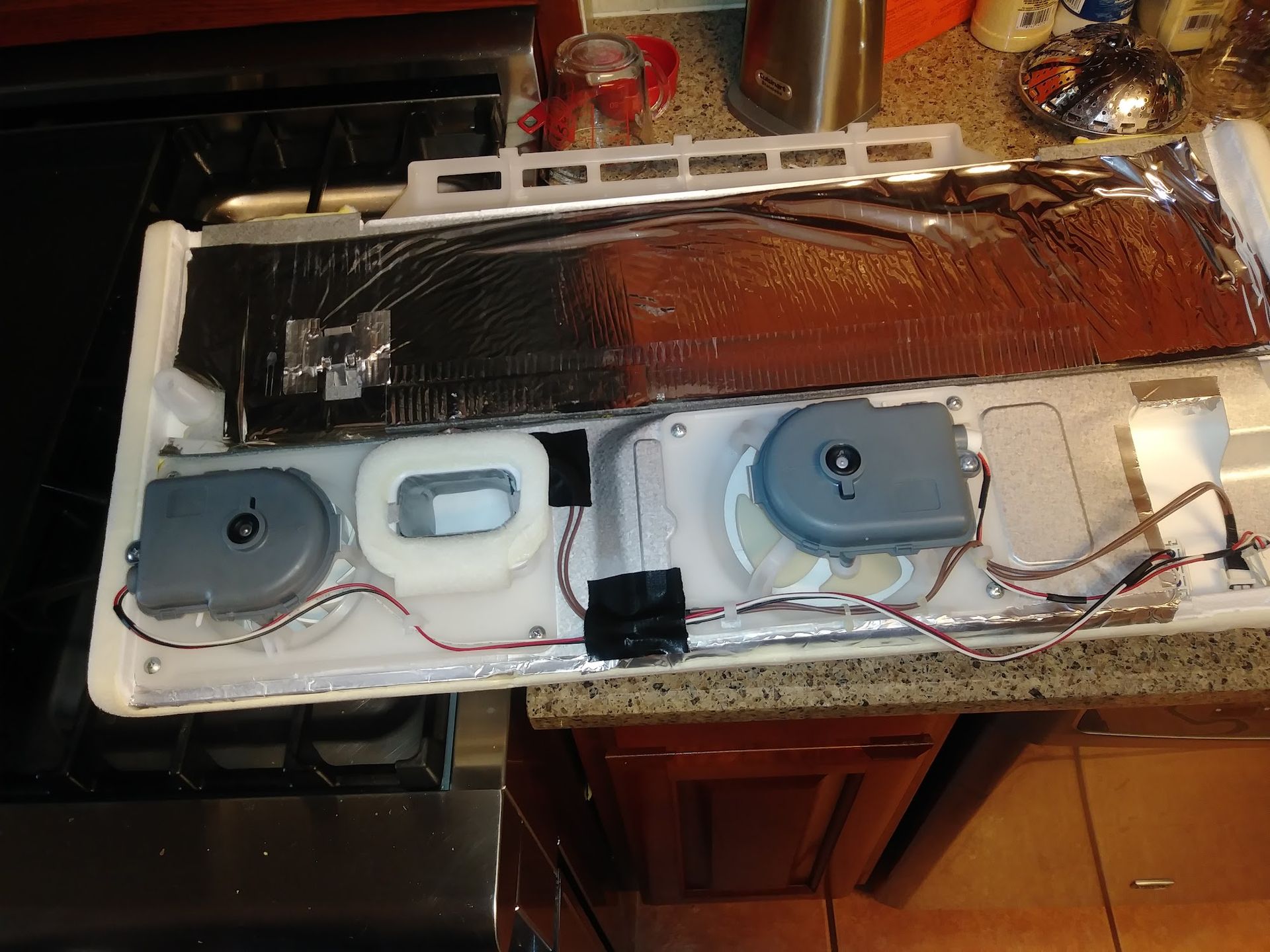 Open refrigerator component with motors, wiring, and black insulation, on a countertop next to a stove.