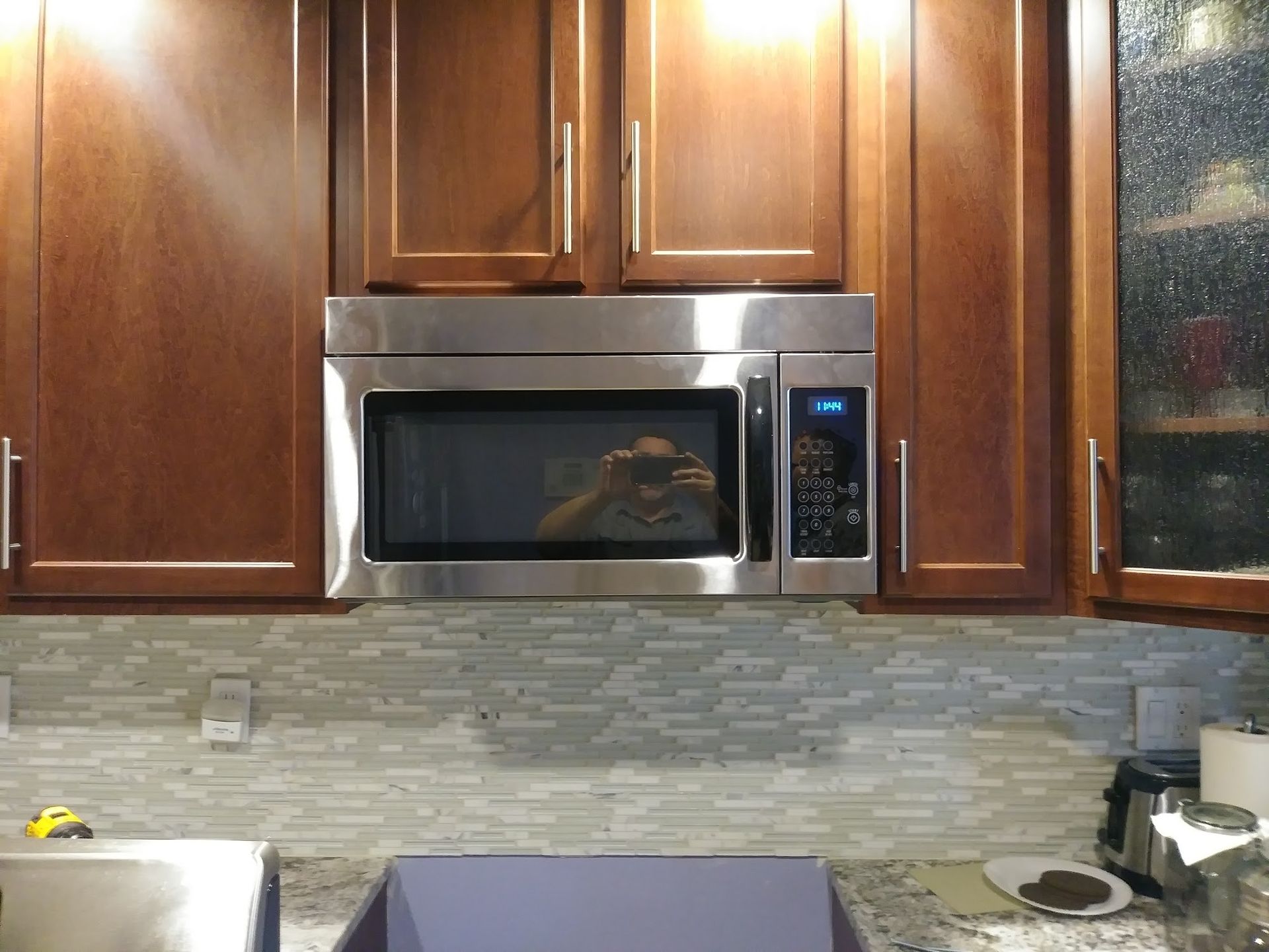 A stainless steel microwave installed under dark brown kitchen cabinets above a gray tiled backsplash.