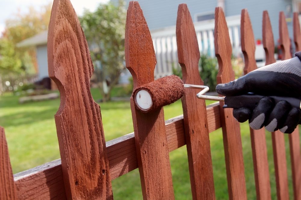 Person in gloves using a roller to apply brown stain to a wooden fence outdoors.