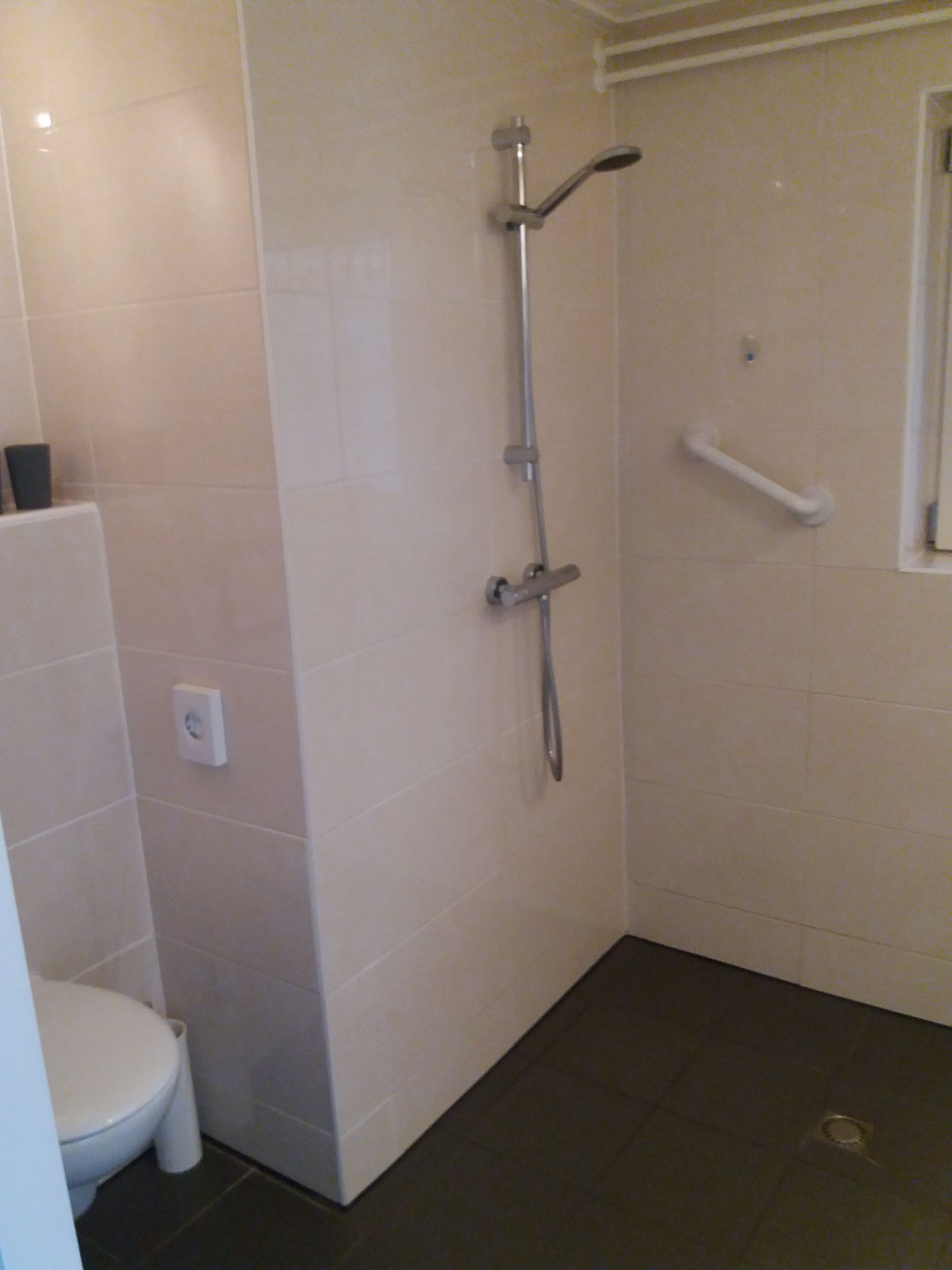 Toegankelijke douchecabine met aan de muur bevestigde handgrepen, handdouche en zitbank in een betegelde badkamer.