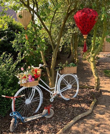 Een witte fiets versierd met bloemen in een zonnige tuin, met rode en gouden hangende lantaarns in de buurt.