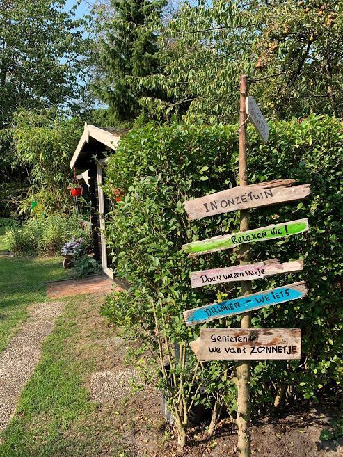 Tuinbordje met kleurrijke pijlen naast een huisje en pad in een zonnige, groene tuin.