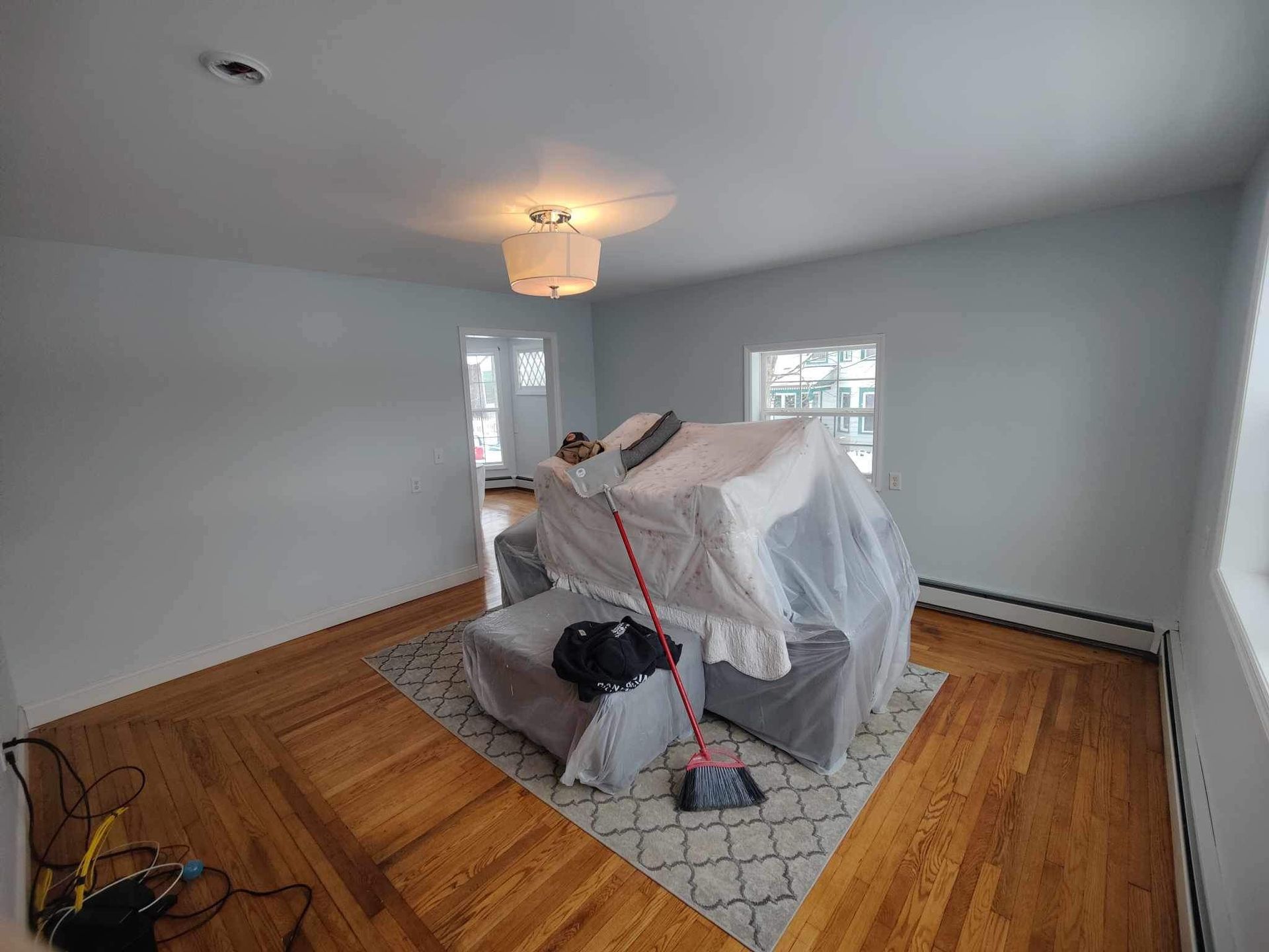 A room with hardwood floors, a covered sofa, and a broom. Sunlight streams through windows.
