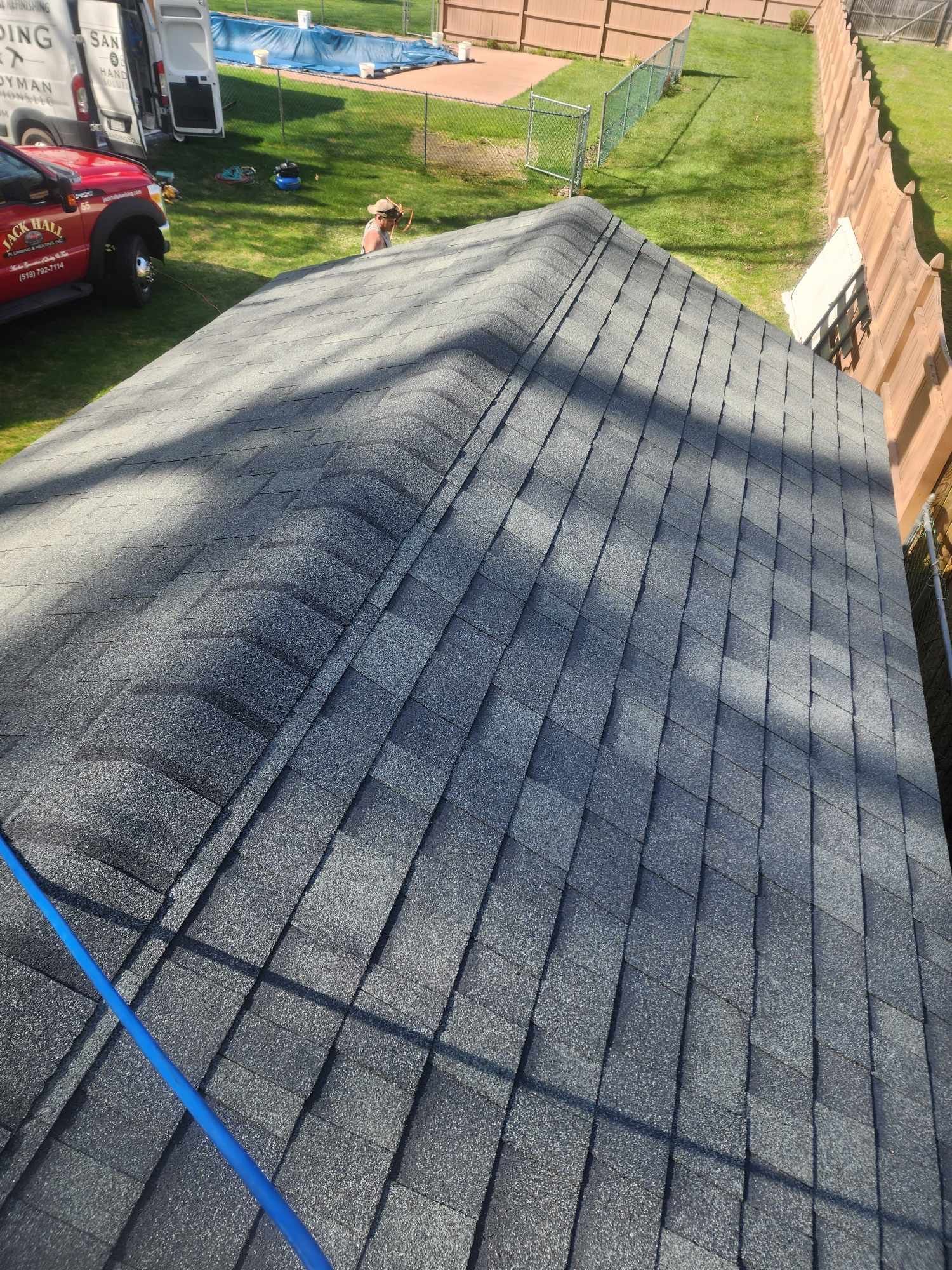 View of a dark shingled roof with a truck and yard in the background on a sunny day.