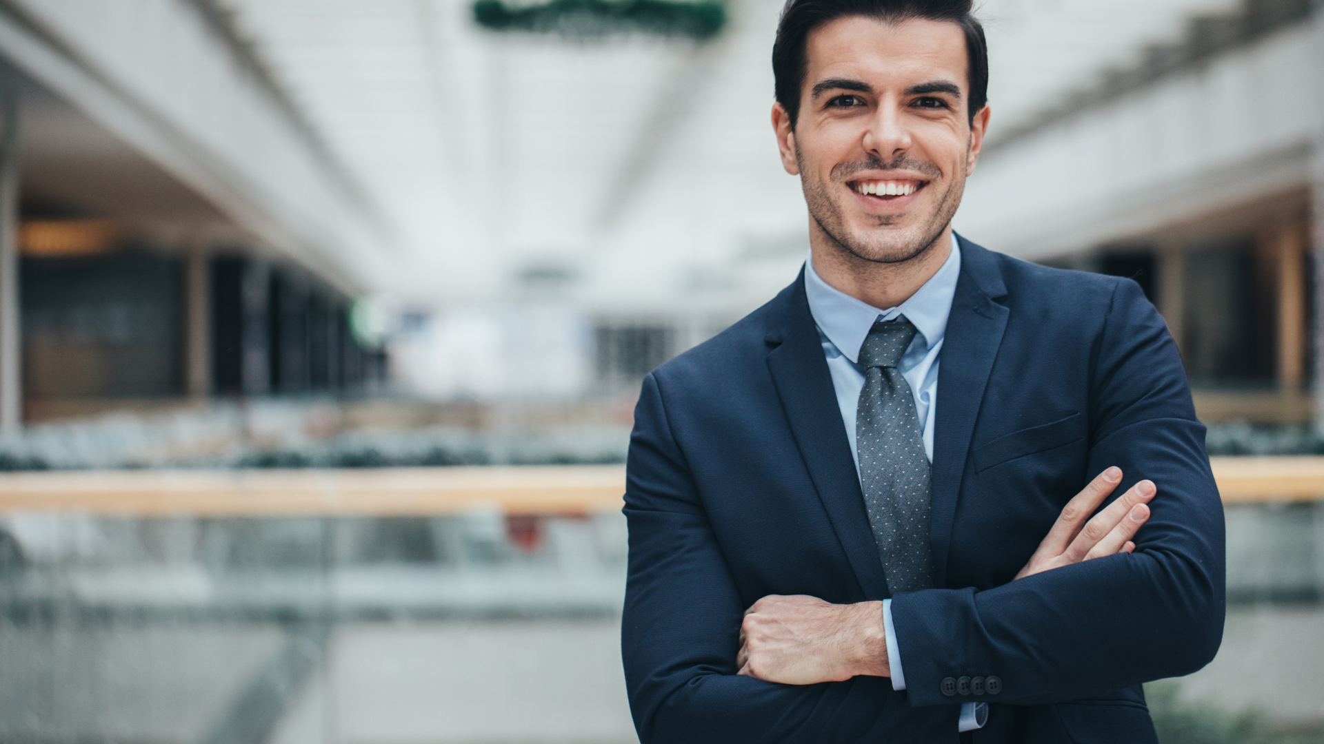 Man in navy suit with arms crossed, smiling, in a bright modern building.
