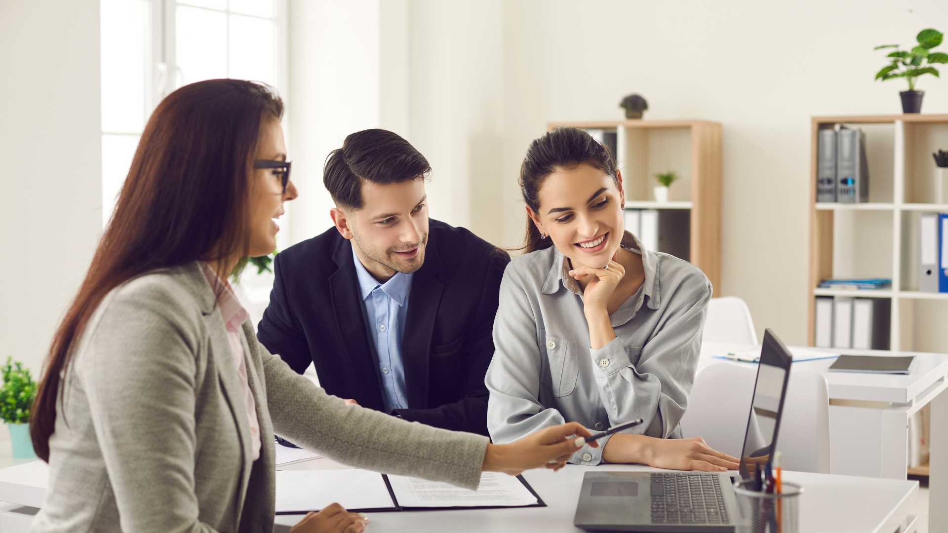 Woman pointing at laptop screen, couple seated, office setting.