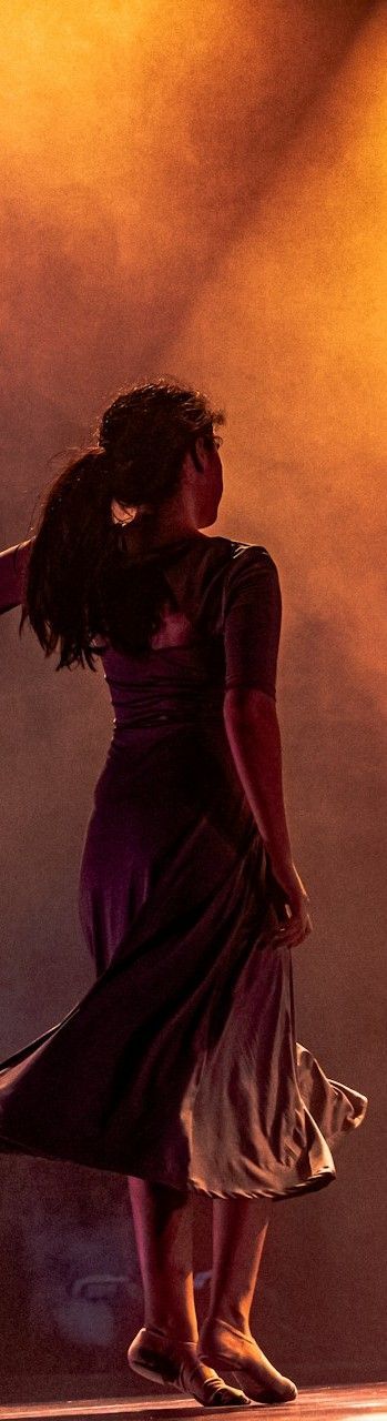 Woman in a dark dress holds up fabric. She turns to her right, in a warm-toned setting.