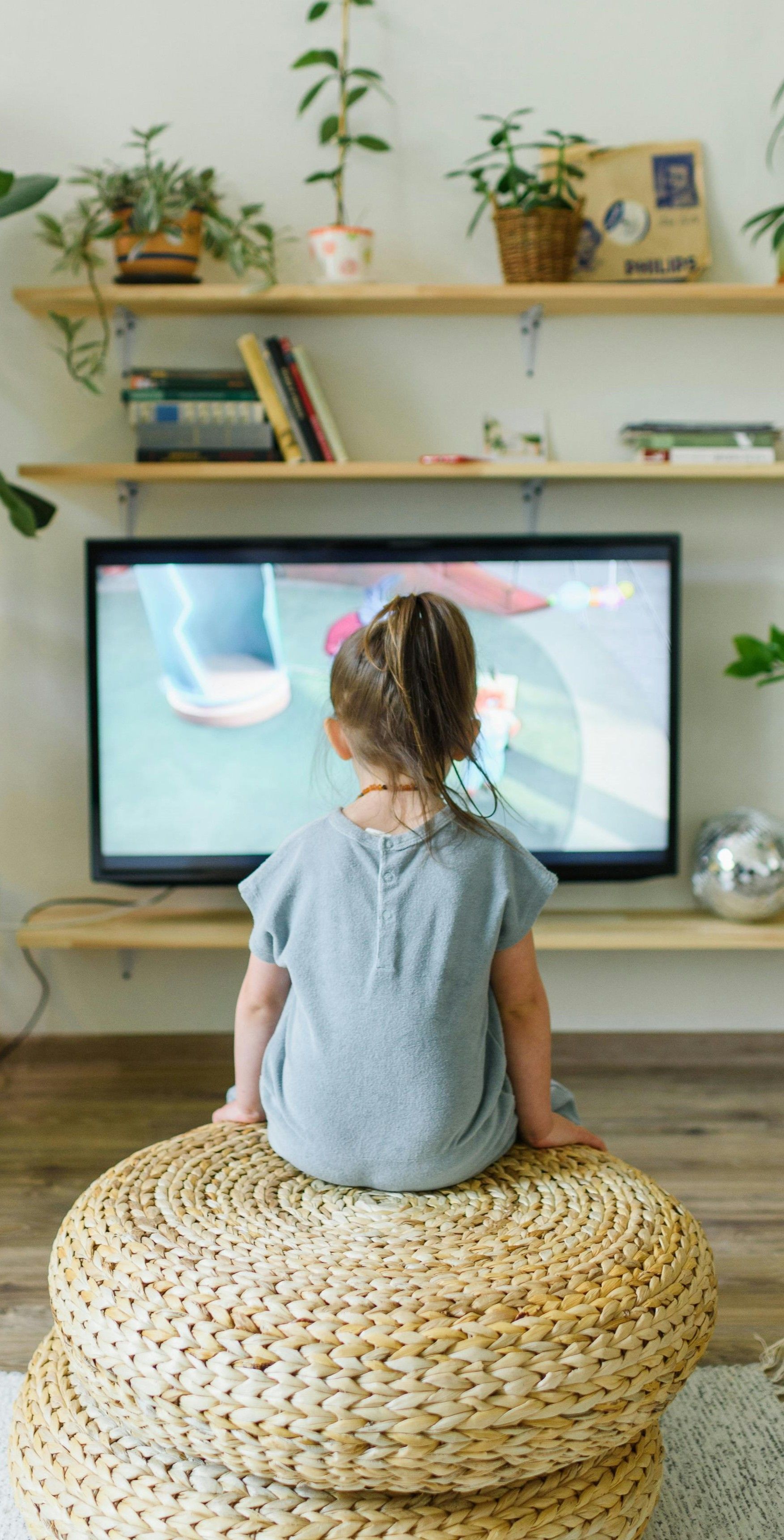 A little girl is sitting on a wicker ottoman watching a television.