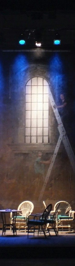 Stage set with a large window illuminated by spotlights, a piano is centred below.