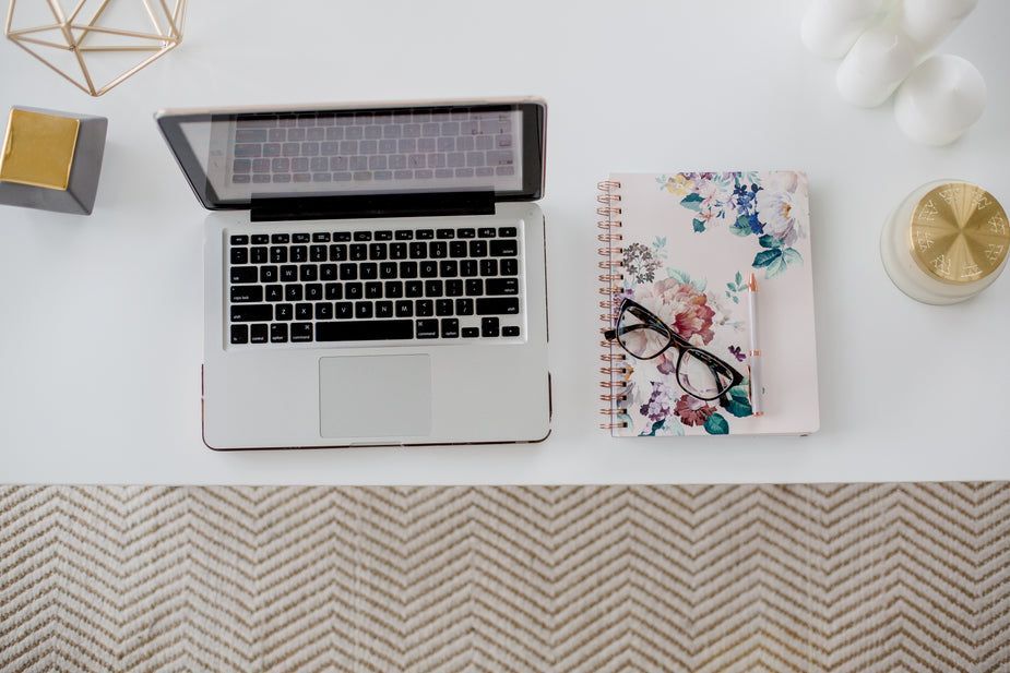 Laptop, floral notebook, glasses, and decor on a white desk with a chevron rug.