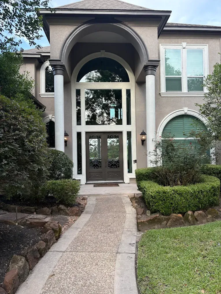 A tan two-story house with a prominent arched entryway, white columns, large glass windows, and a pebble walkway.