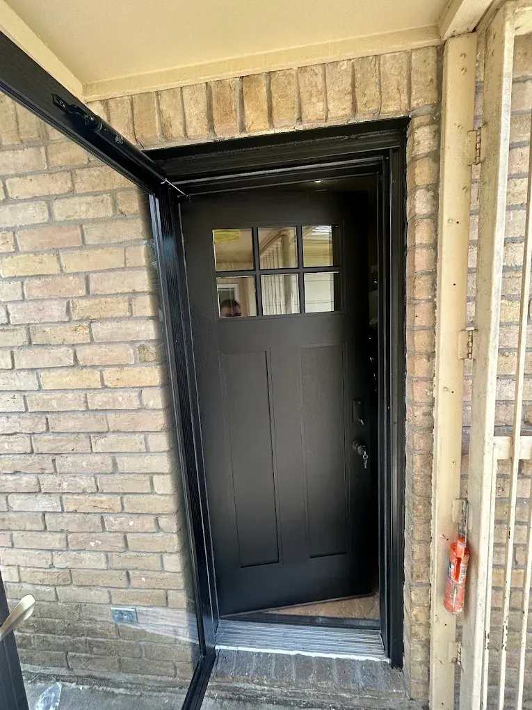 A black entry door with a six-pane window at the top, set in a light-colored brick wall under a covered porch.