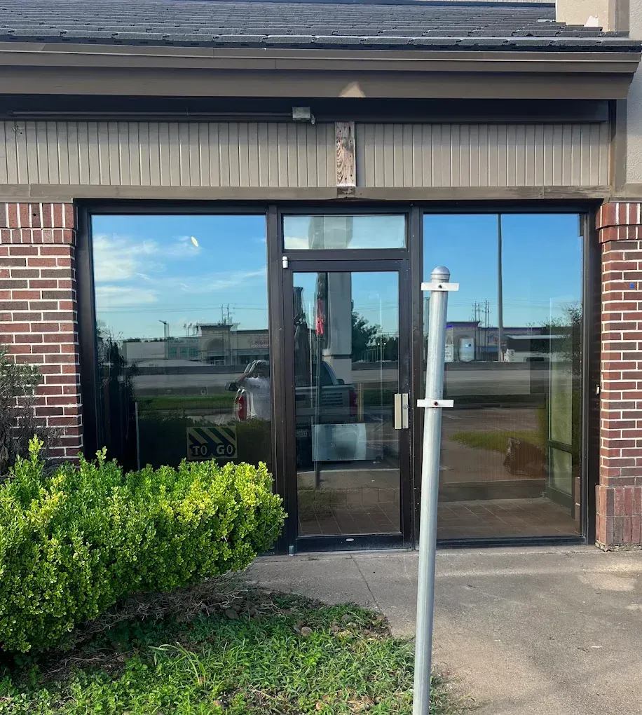 A glass entry door with dark metal frames, flanked by large windows on a brick building with a gray awning and green bush.