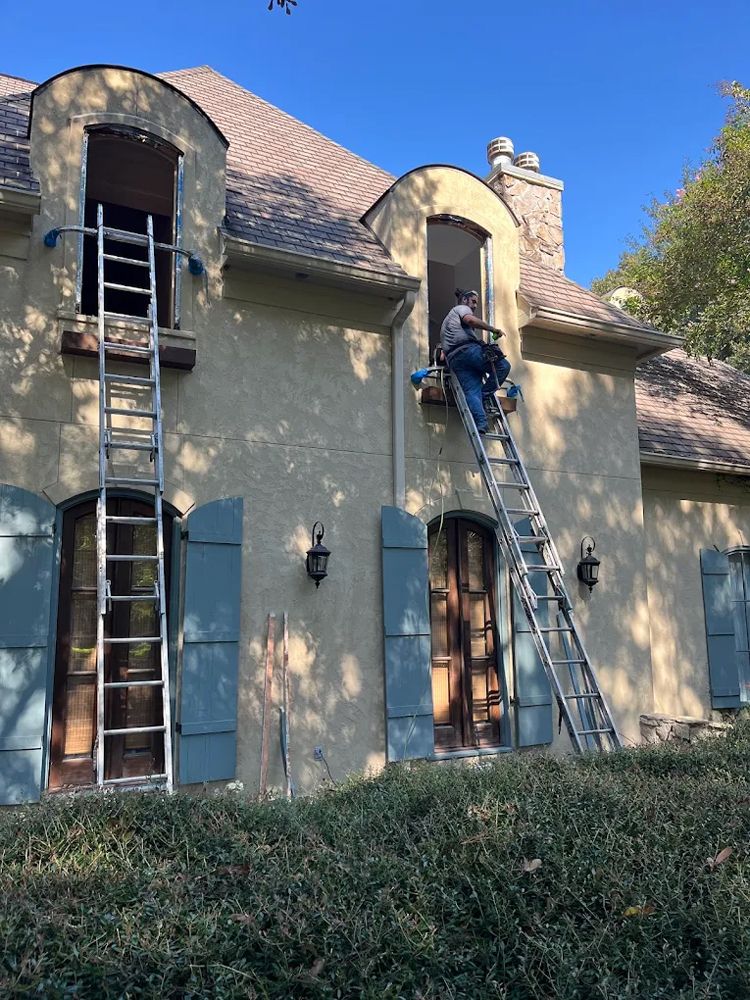 A person on a ladder entering an open second-story window of a tan house with blue shutters on a sunny day.