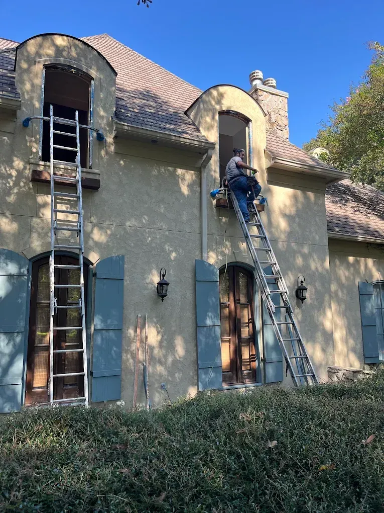 A person on a ladder repairs a second-story window of a tan stucco house with blue shutters on a sunny day.