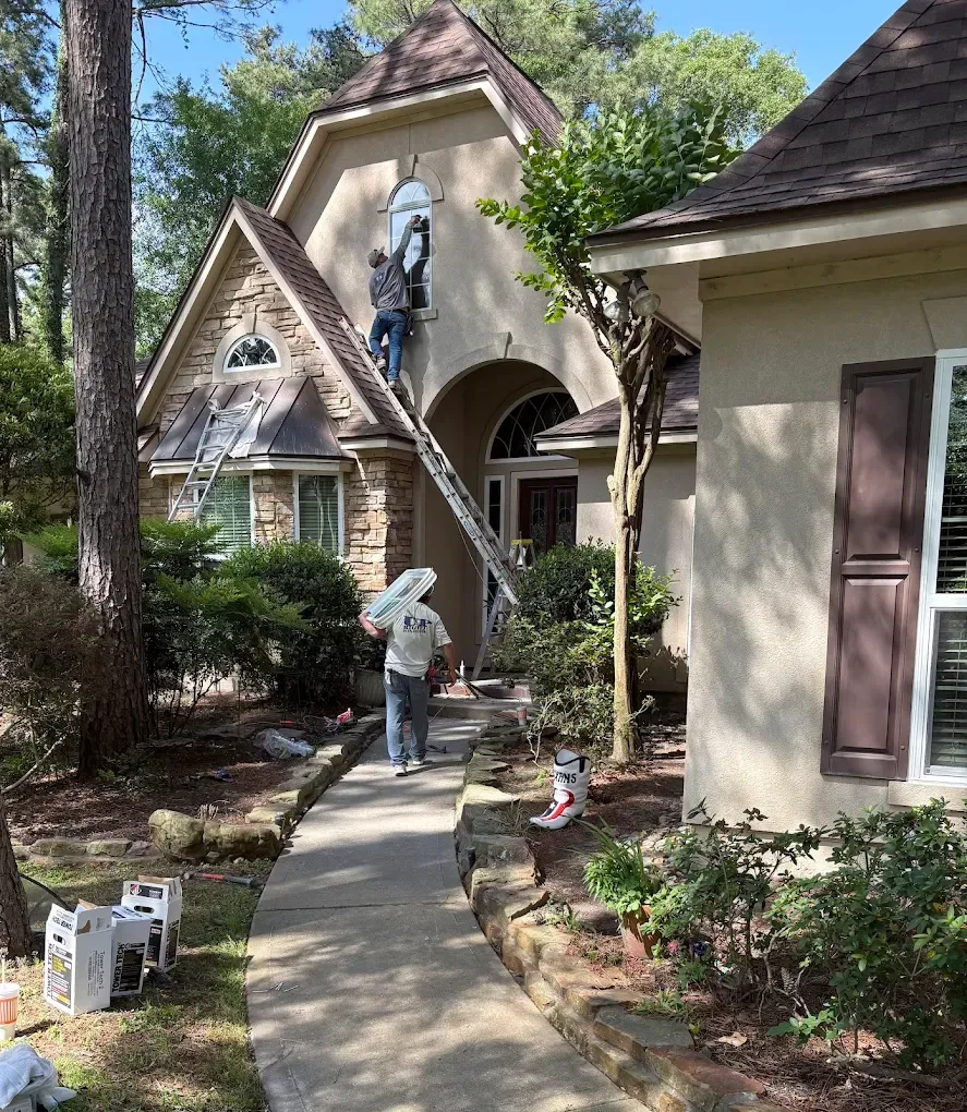 Workers use a ladder to install an arched window on the upper exterior of a beige stone and stucco house.
