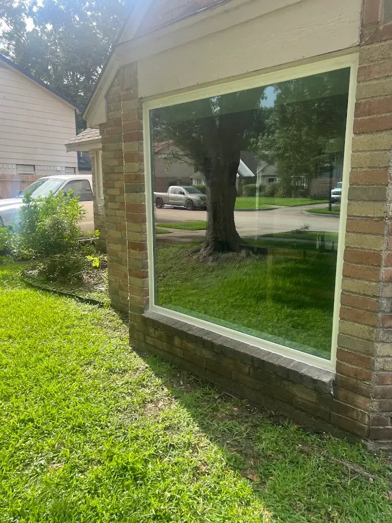 A large square window on a brick exterior wall reflecting a tree, a parked car, and a residential street.